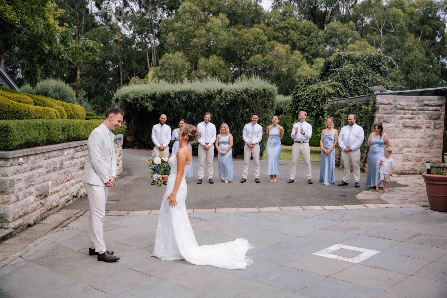 Bride leans in to kiss groom during outdoor first look with wedding party watching on stone patio.
