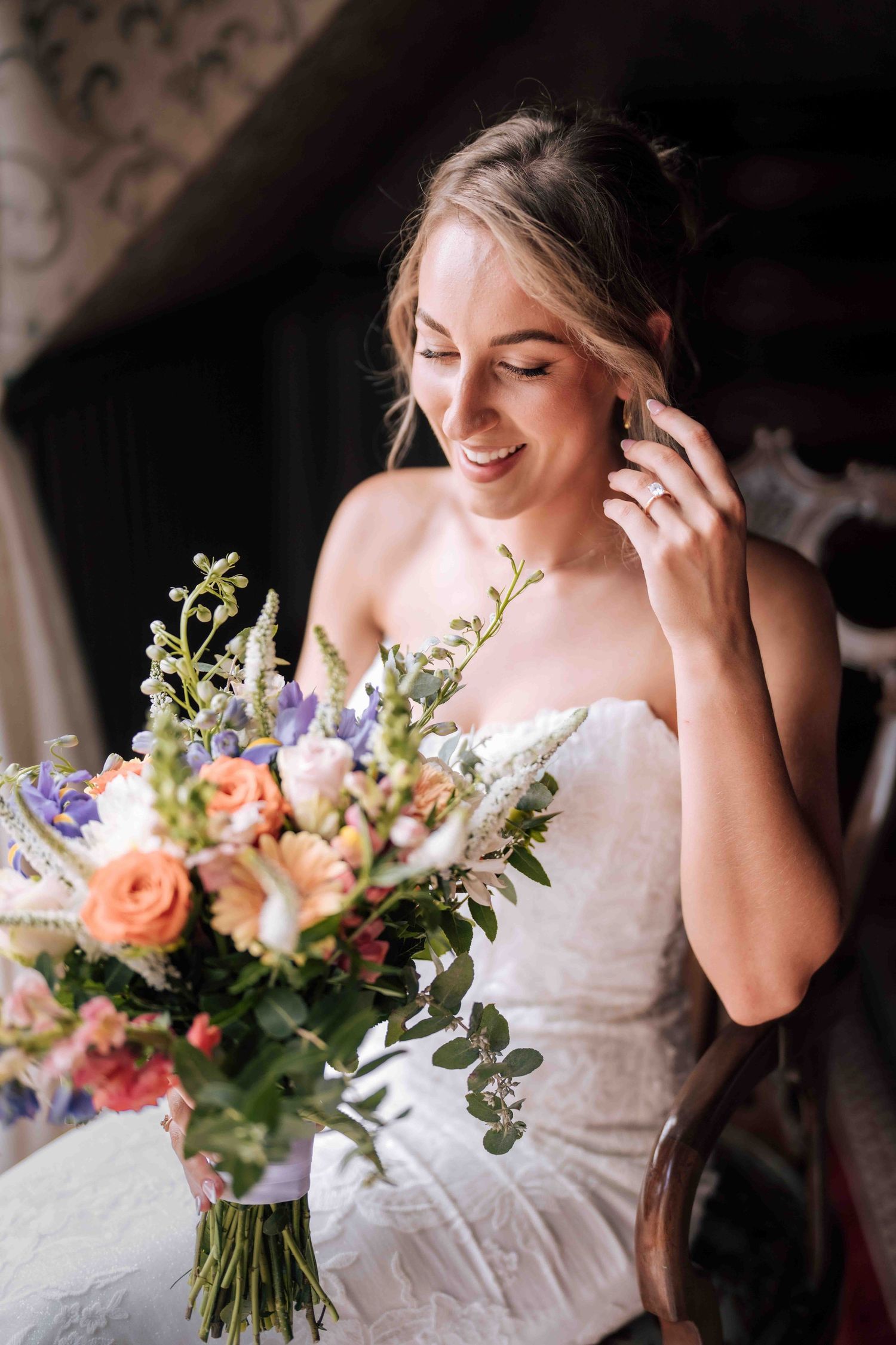A smiling bride in a strapless white lace gown adjusts her earring while holding a vibrant floral bouquet.