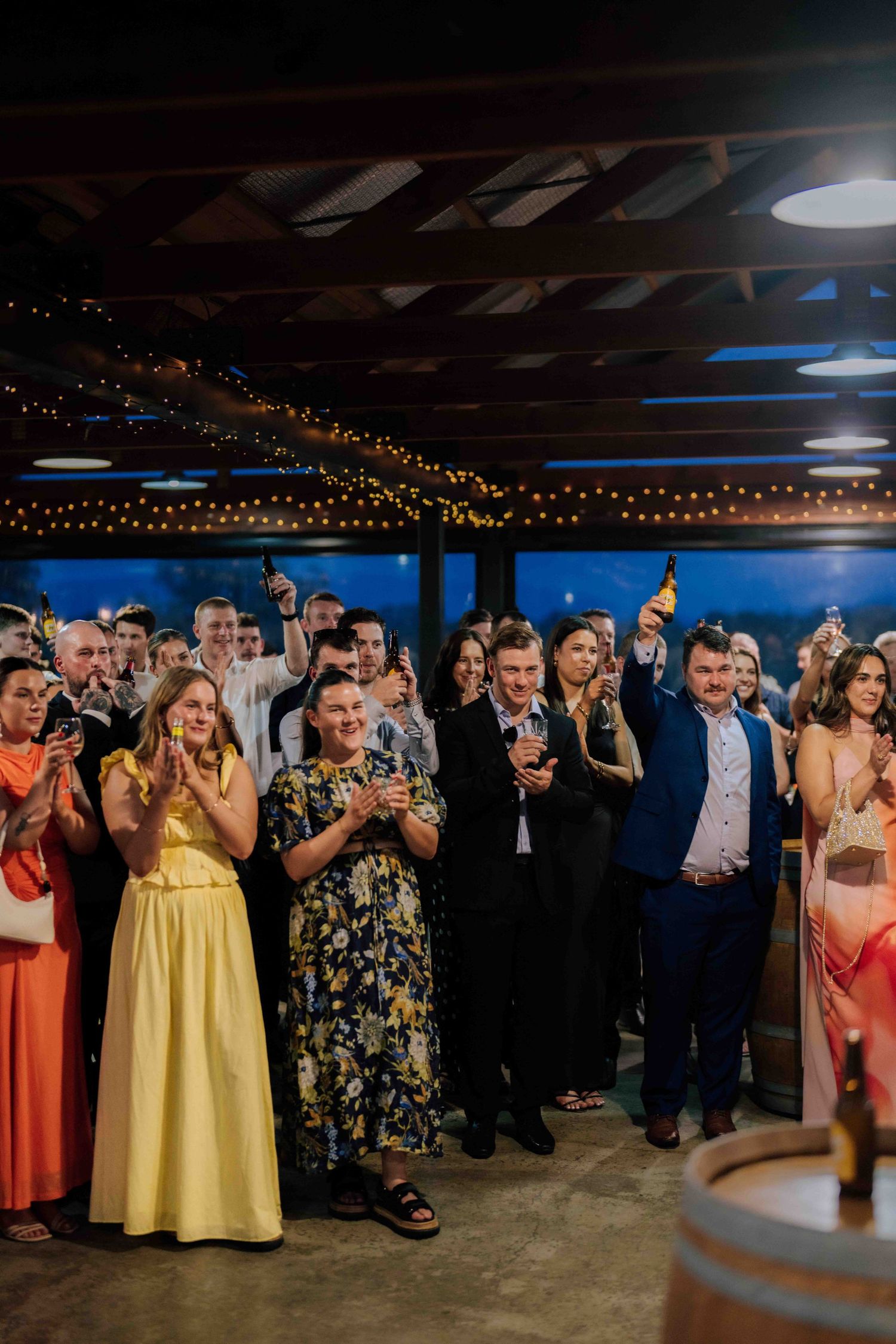 Wedding guests clapping and raising glasses in celebration during indoor reception with string lights.