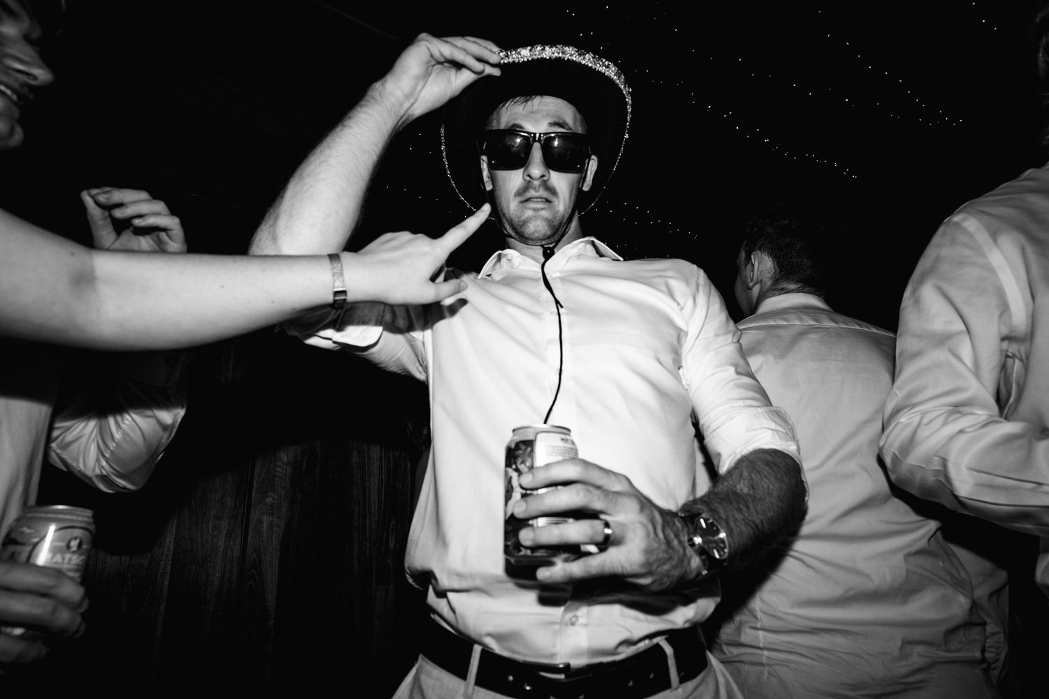 Man in sunglasses dancing with beer at lively black and white wedding reception party.