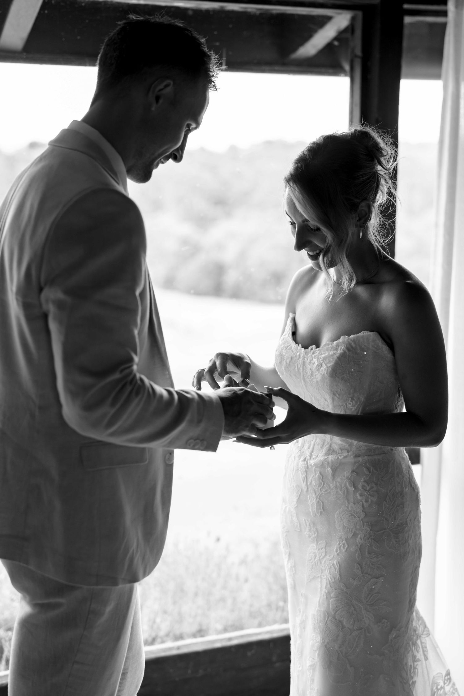 Black and white silhouette of bride and groom exchanging wedding rings by a sunlit window in a barn venue.