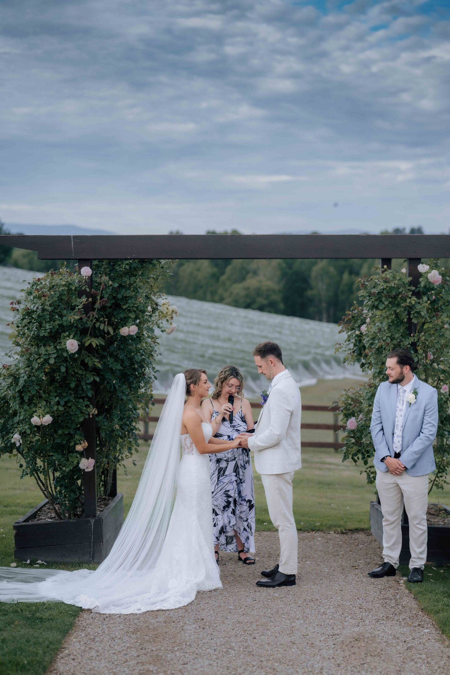 Bride and groom exchange vows at floral arch with celebrant and groomsman at vineyard wedding.