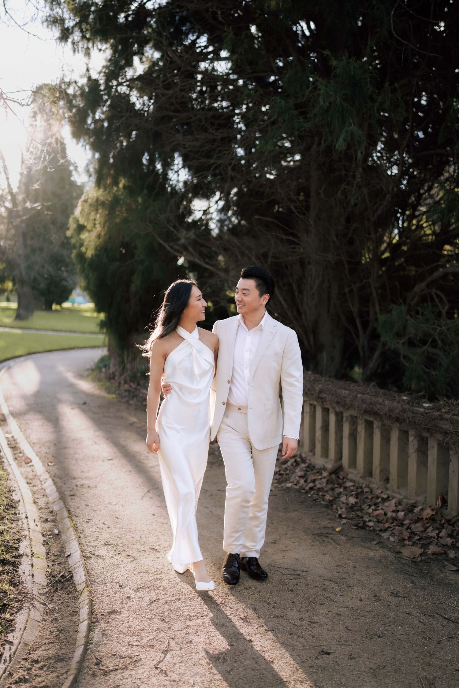 Couple in white outfits walking hand in hand along a sunlit tree-lined path during a romantic outdoor photoshoot.