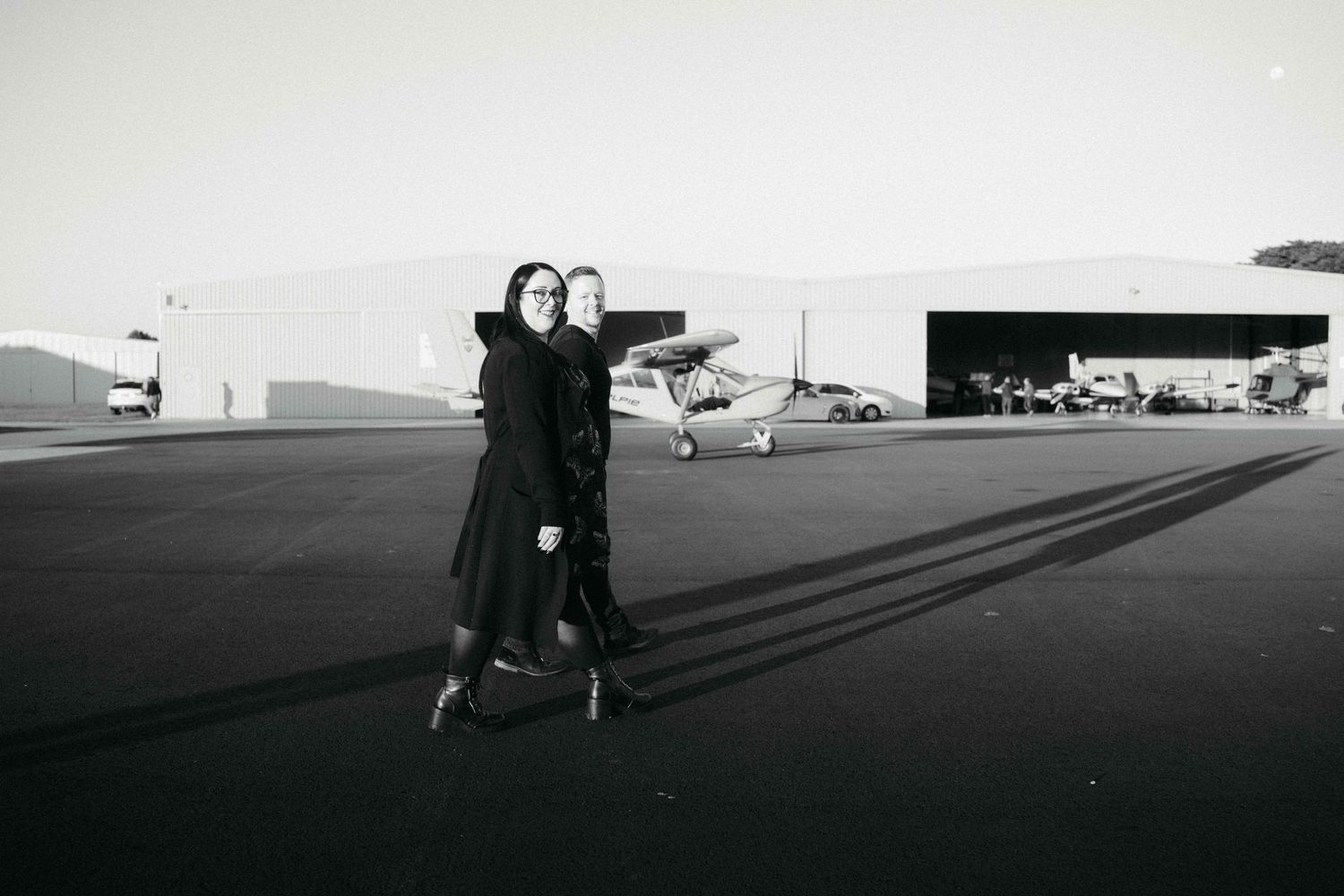 Two people embracing in black outfits on an airport tarmac with small aircraft and hangar in background, black and white photo.