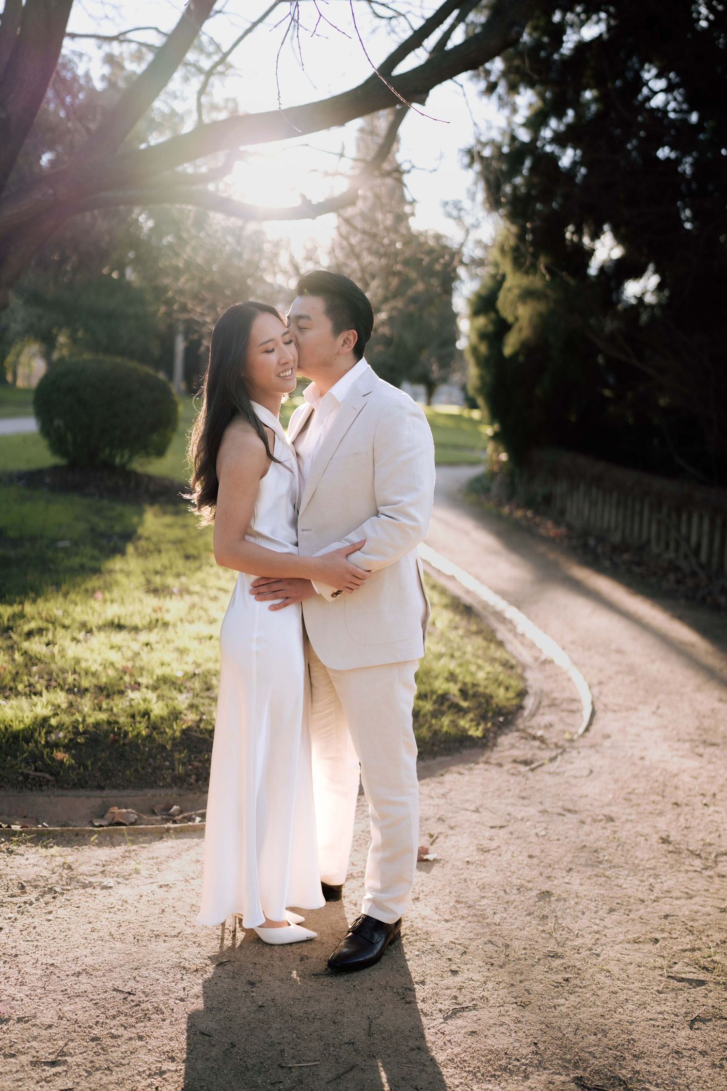 Couple in white attire embracing on a sunlit park path, sharing a romantic moment amid lush greenery.