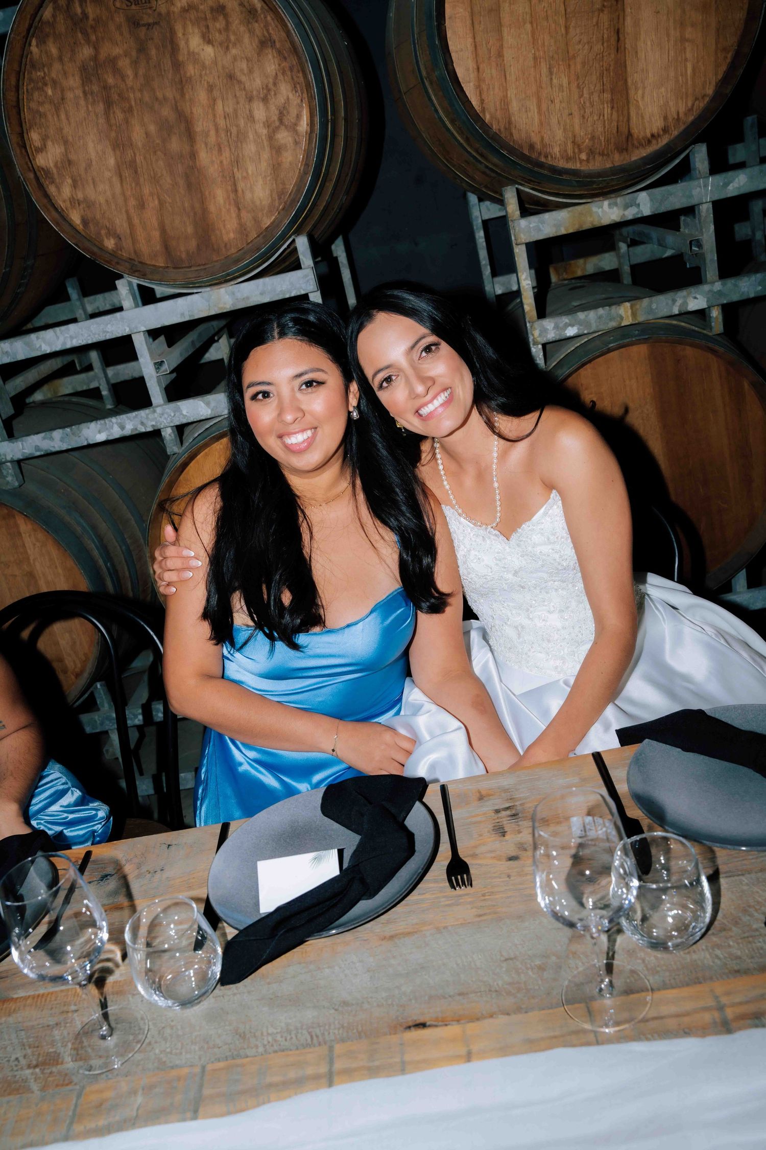 Bride poses with bridesmaid in blue satin dress seated at rustic table near wine barrels.
