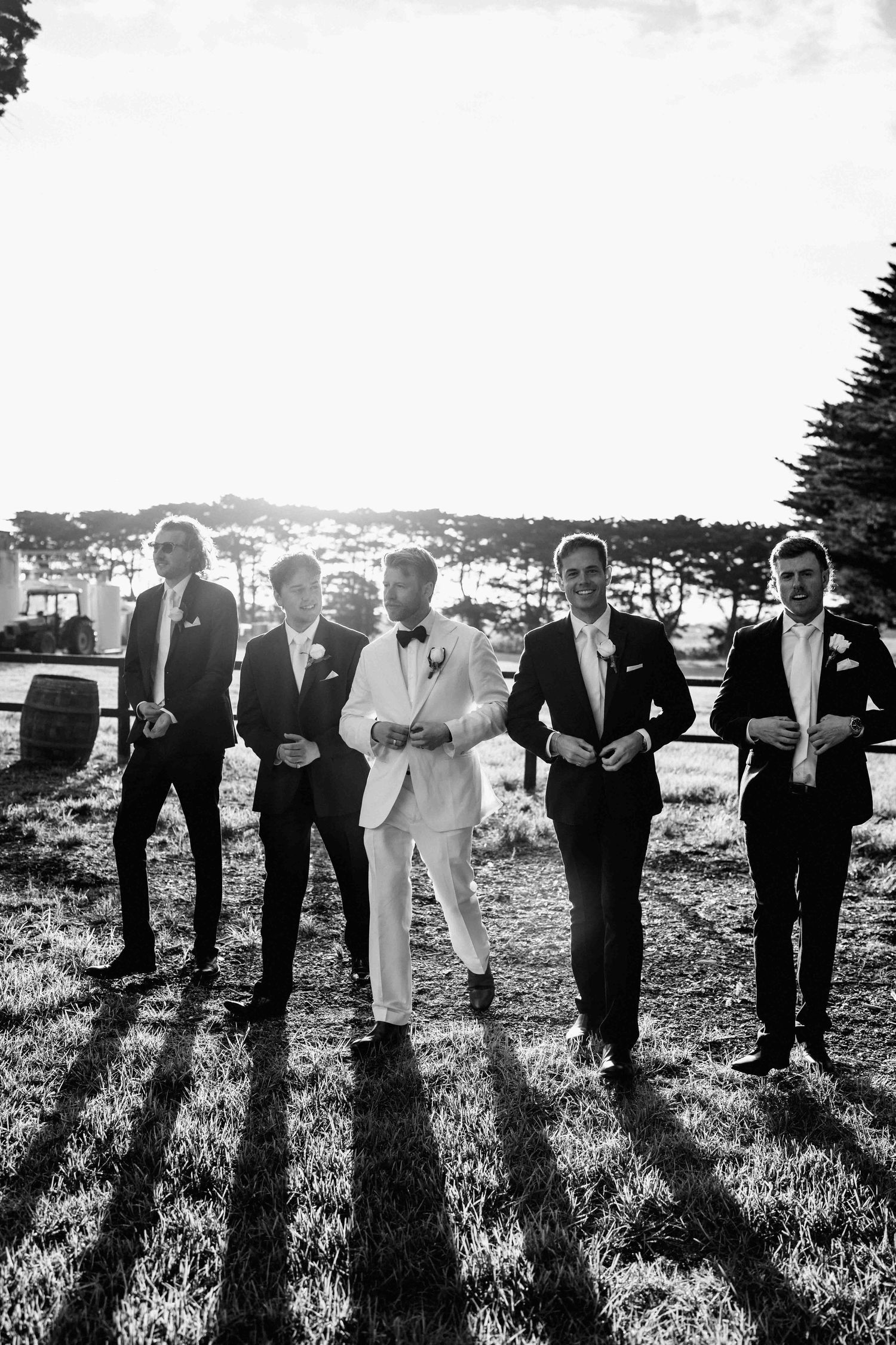 Black and white photo of groom in white suit walking with four groomsmen in dark suits across a sunlit field.