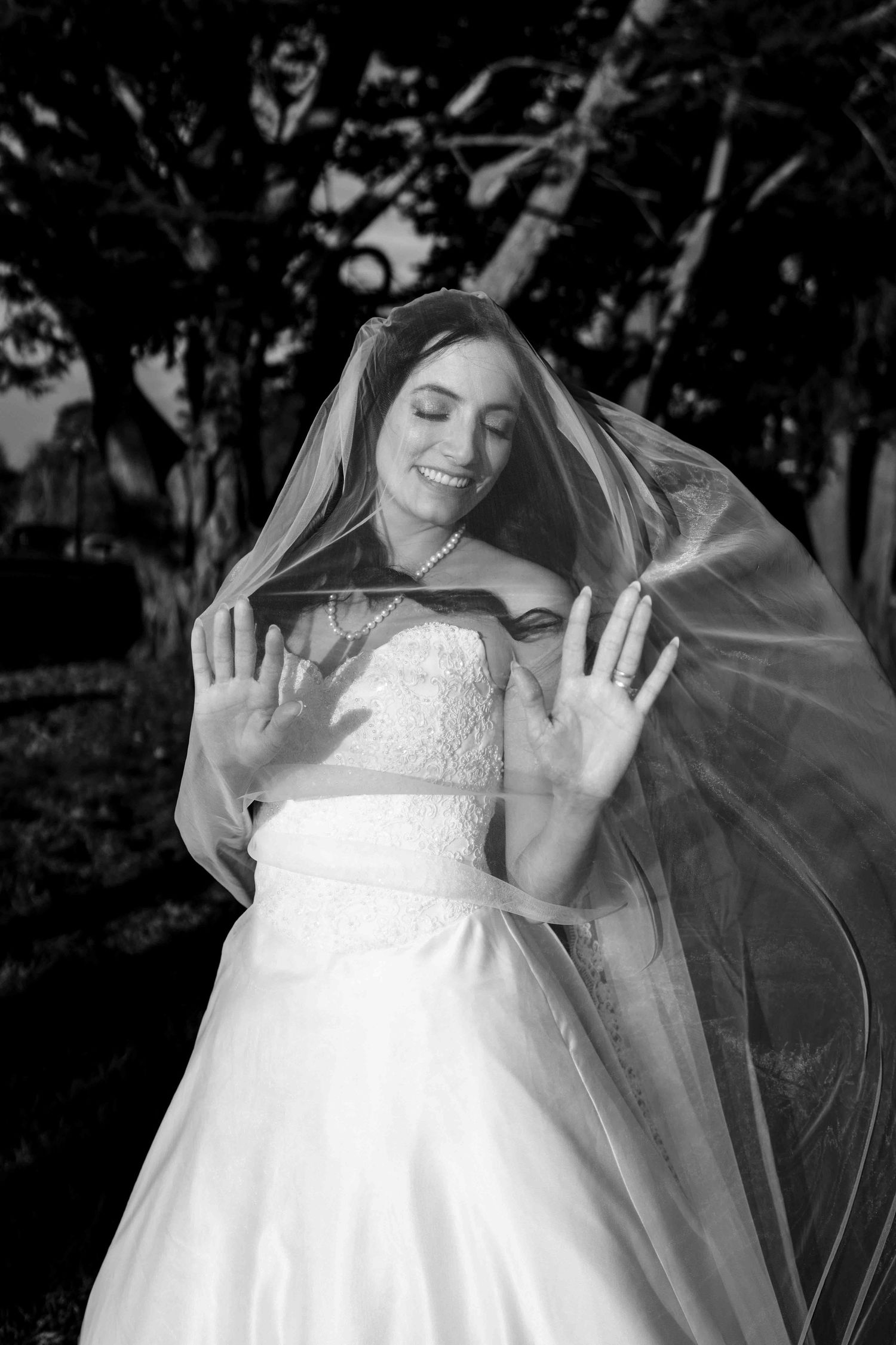 Smiling bride with veil draped over her face, hands raised, black and white bridal portrait outdoors.