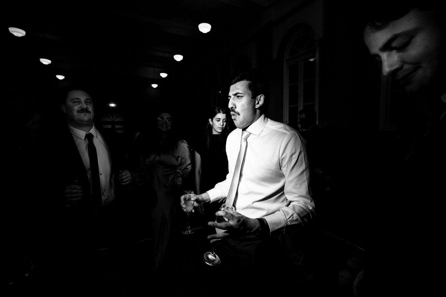 Black and white candid photo of wedding guests socializing in dimly lit reception venue.