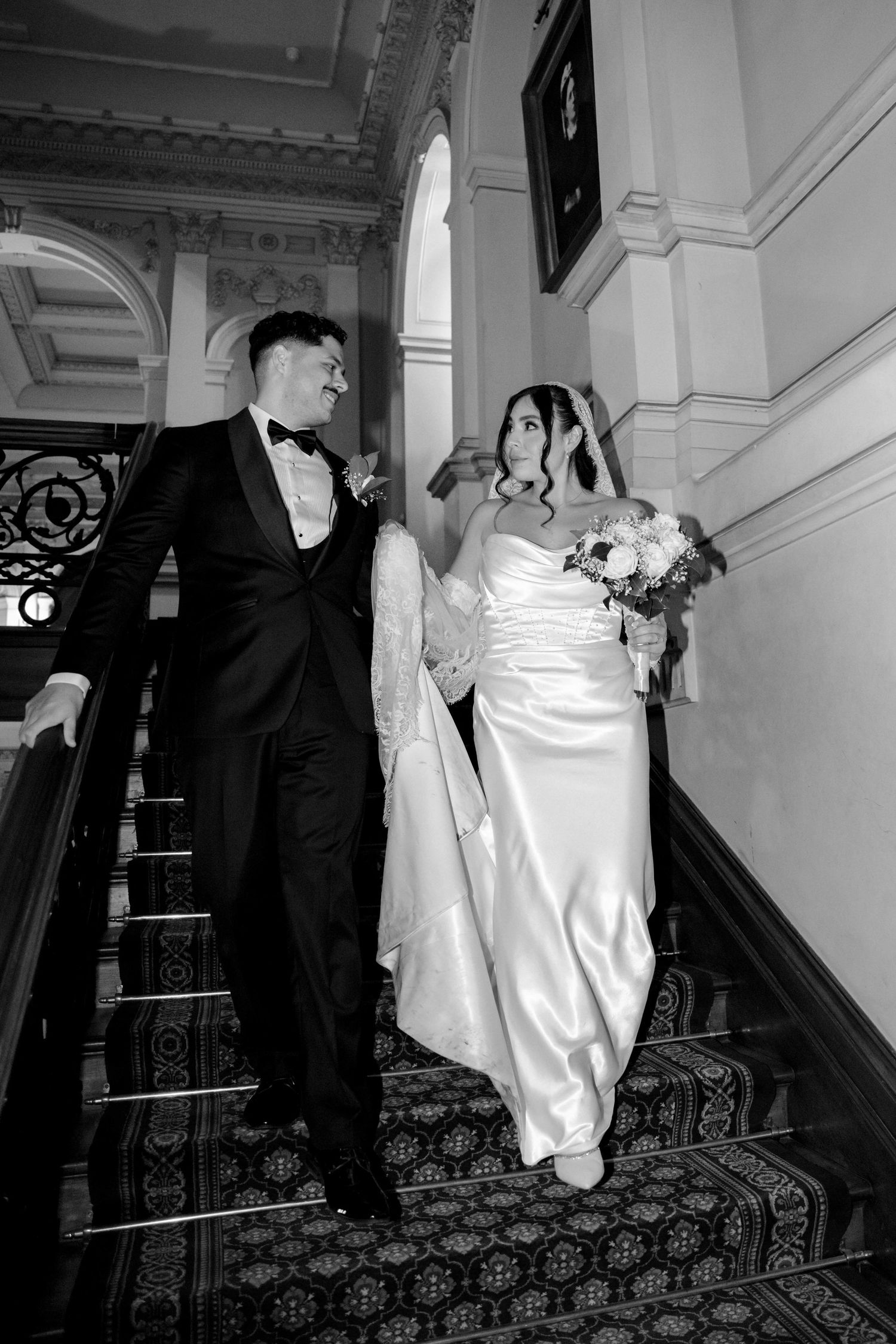 Black and white photo of bride and groom descending elegant staircase together.