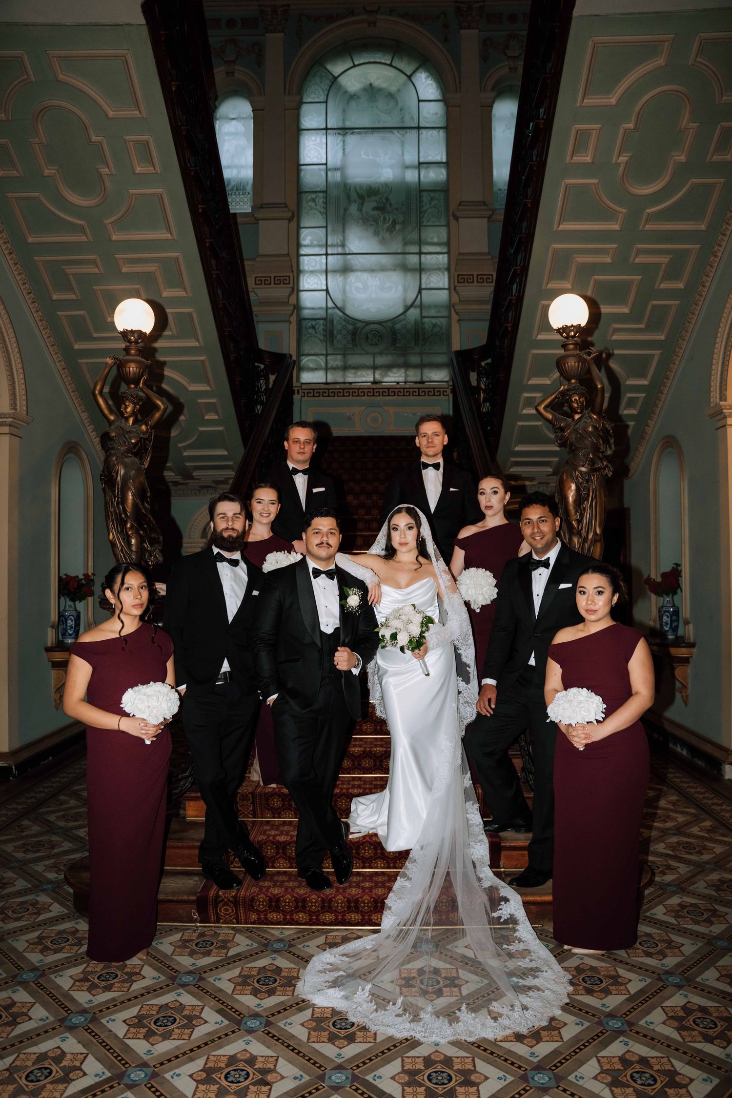 Wedding party in black tuxedos and burgundy gowns posing on grand staircase with stained glass window.