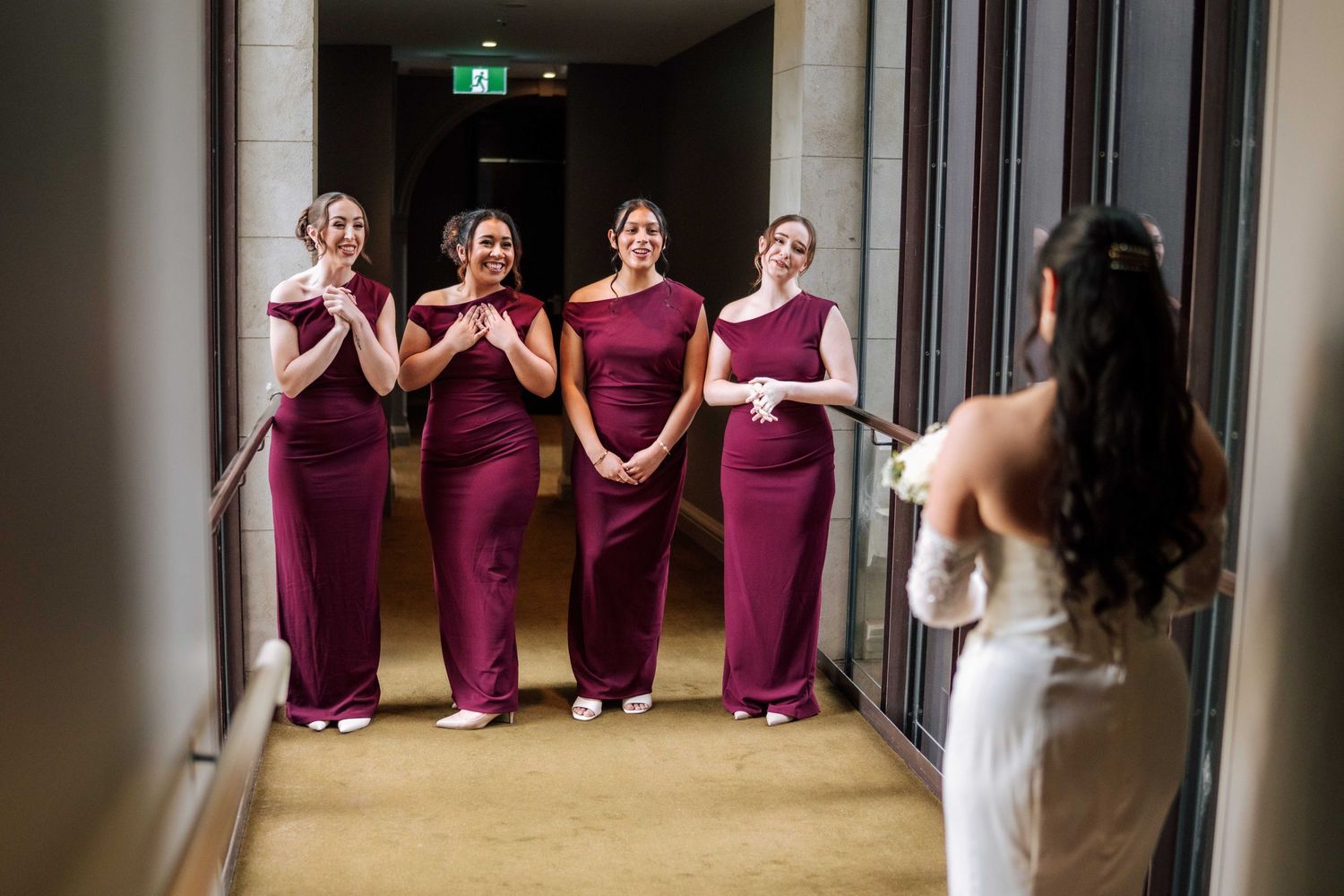 Bride reveals wedding dress to emotional bridesmaids in burgundy gowns standing in a hotel corridor.