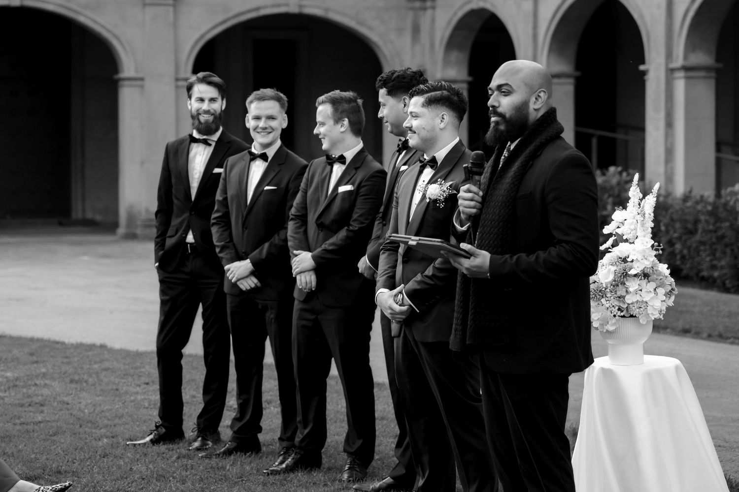 Groom and groomsmen in black tuxedos standing at outdoor wedding ceremony altar.