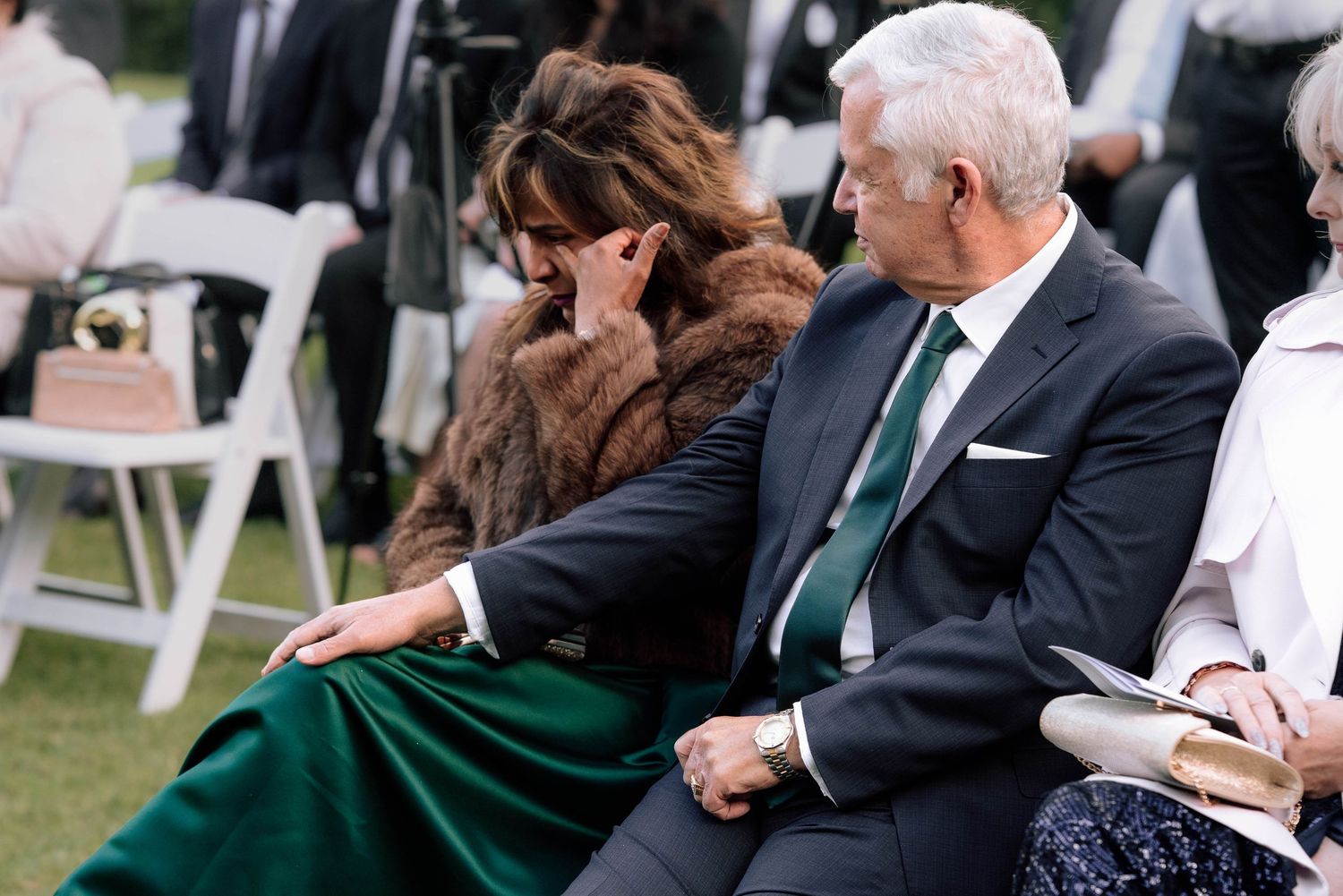 Emotional wedding guests sitting in white chairs during outdoor ceremony, woman wiping tears.