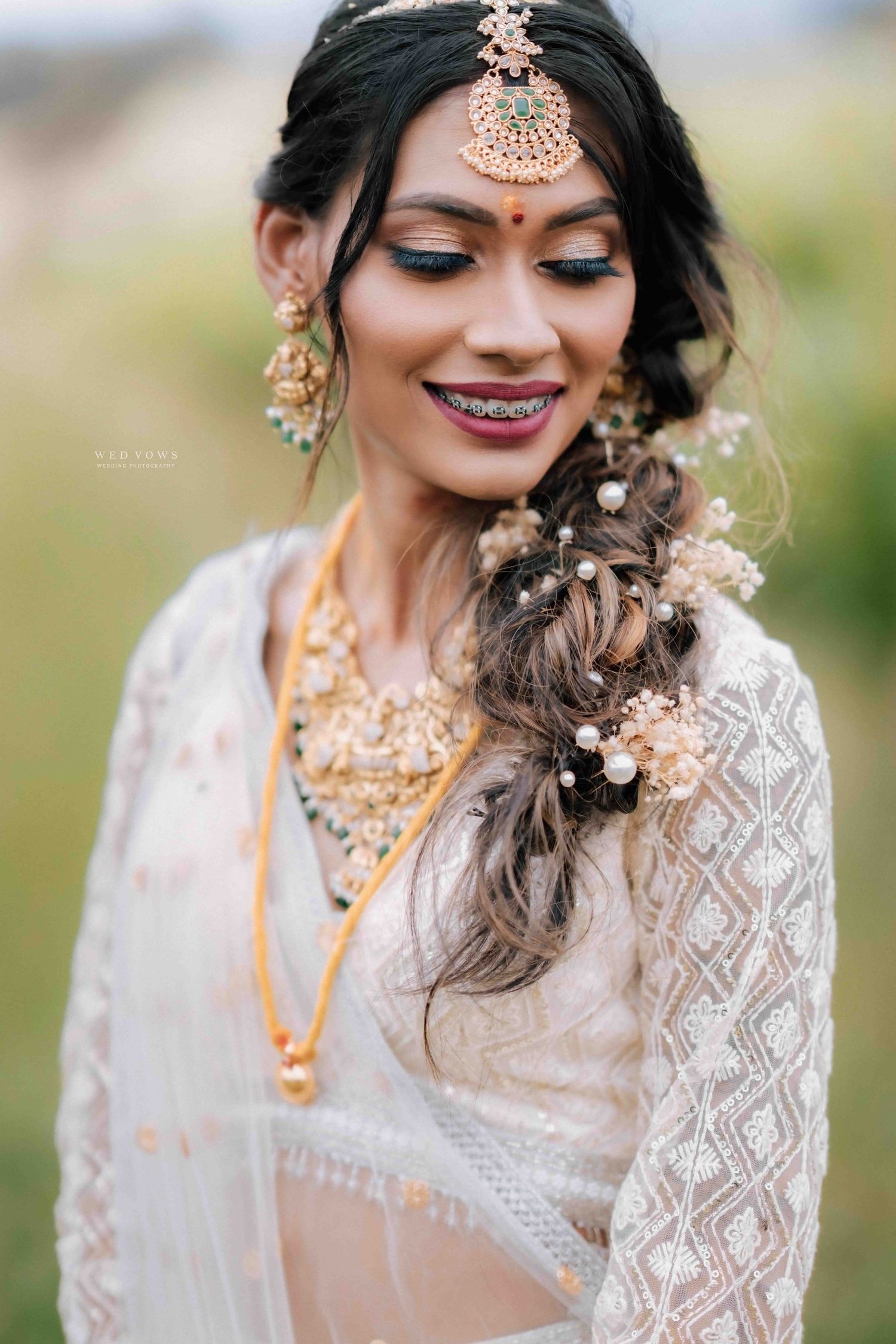 Beautiful Indian bride smiles wearing maang tikka, floral hair accessories, and gold jewelry.