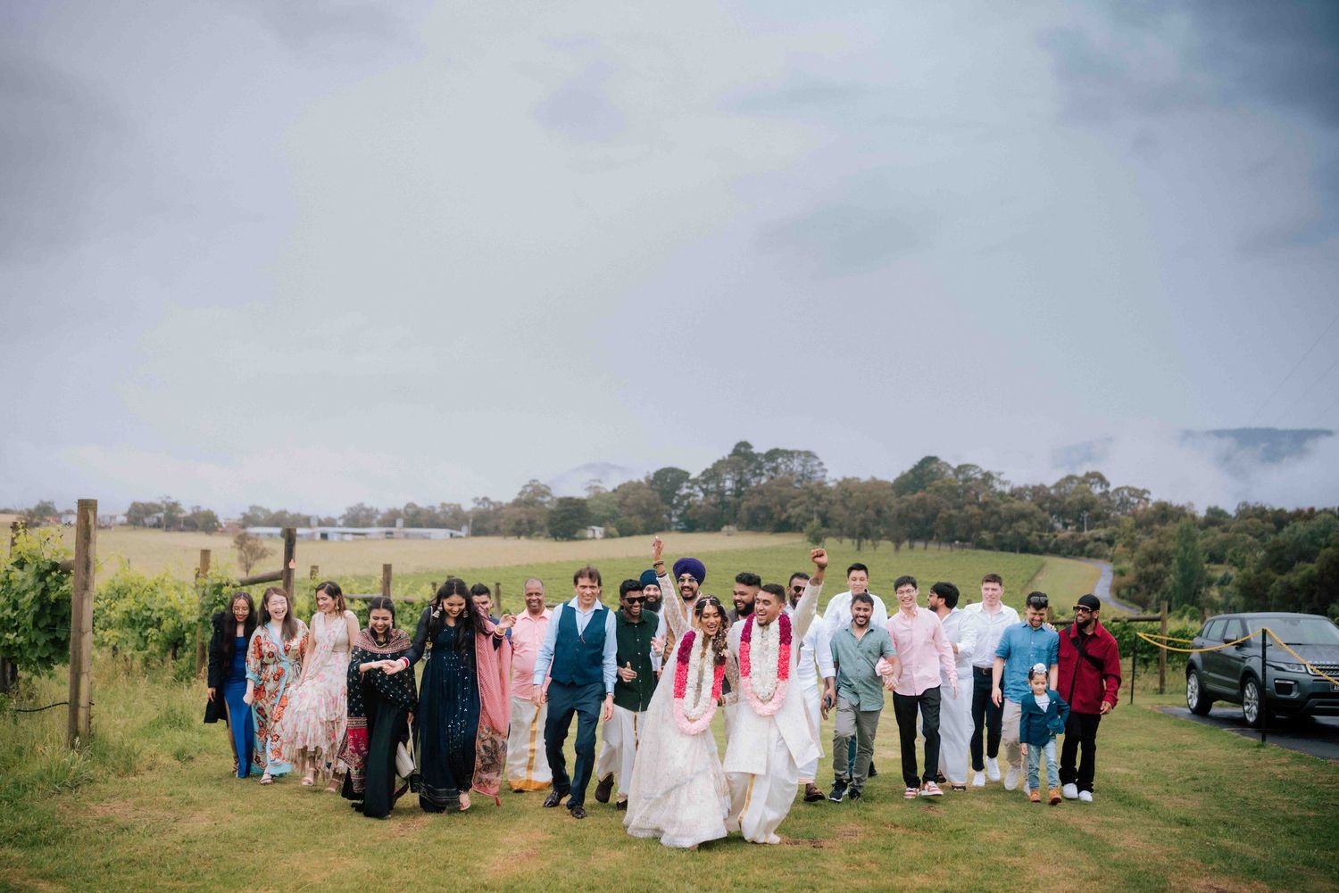 Wedding party walking together through a scenic vineyard with rolling green hills and misty mountains in the background.
