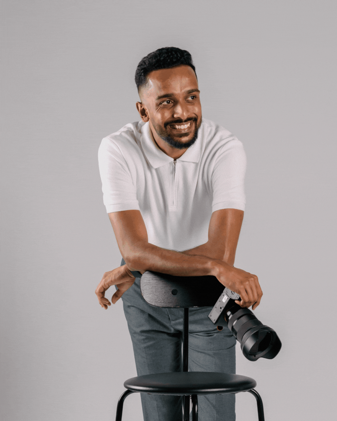 Smiling male photographer in white polo leaning on bar stool with professional camera, gray background.