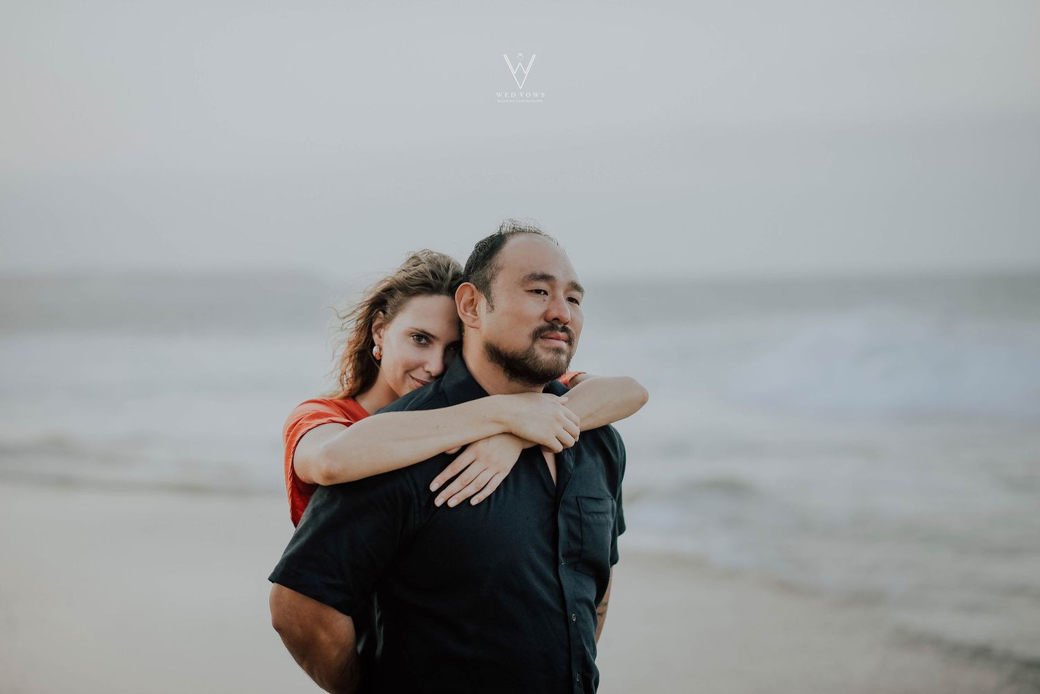Woman embracing man from behind on a beach with ocean waves in the background.