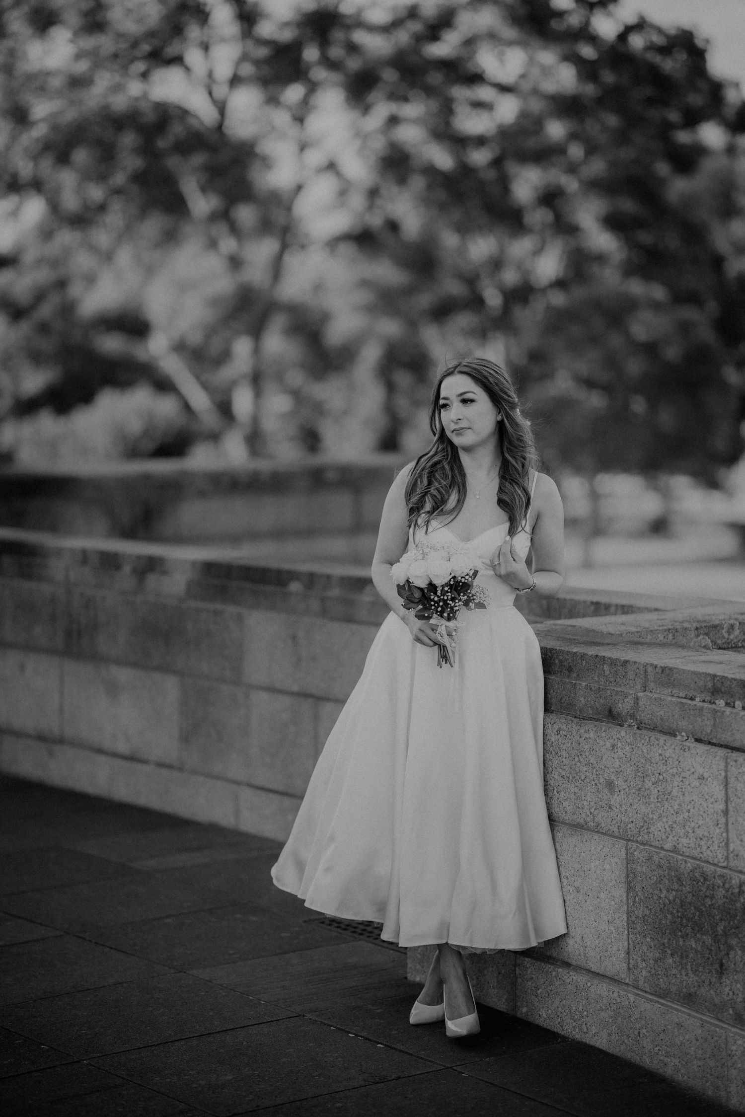 Elegant bride in tea-length white dress holds bouquet while leaning against stone wall in black and white photo.