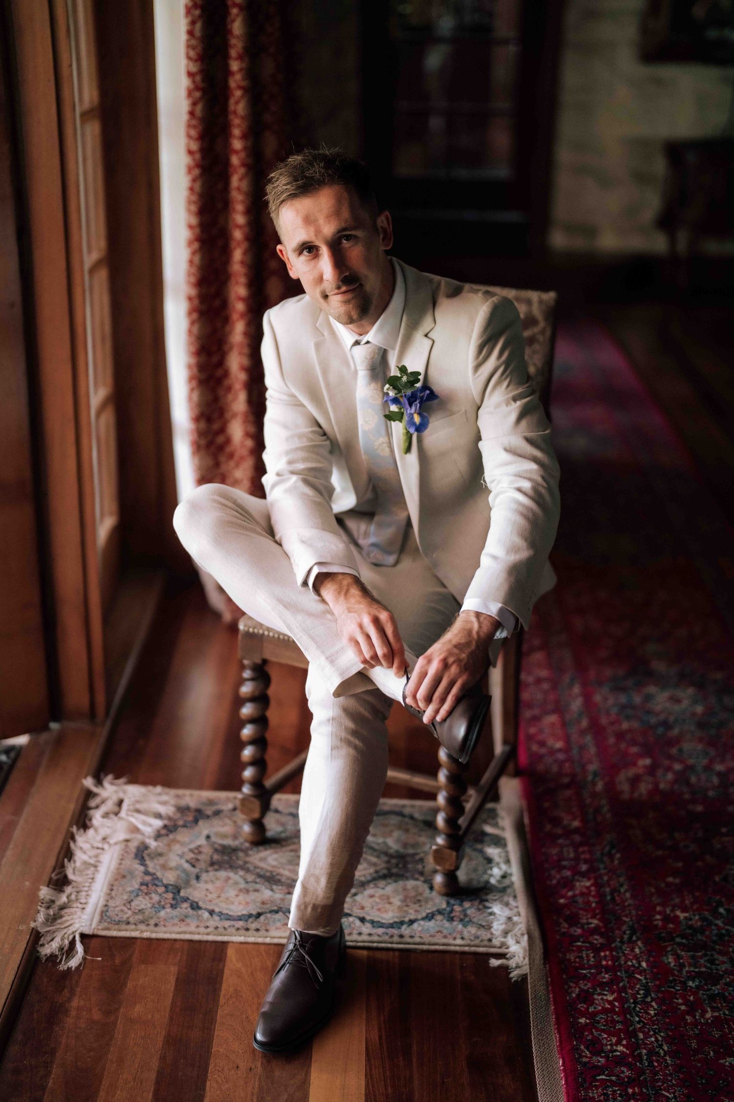 Elegant groom in white linen suit with blue boutonniere seated on vintage chair indoors.