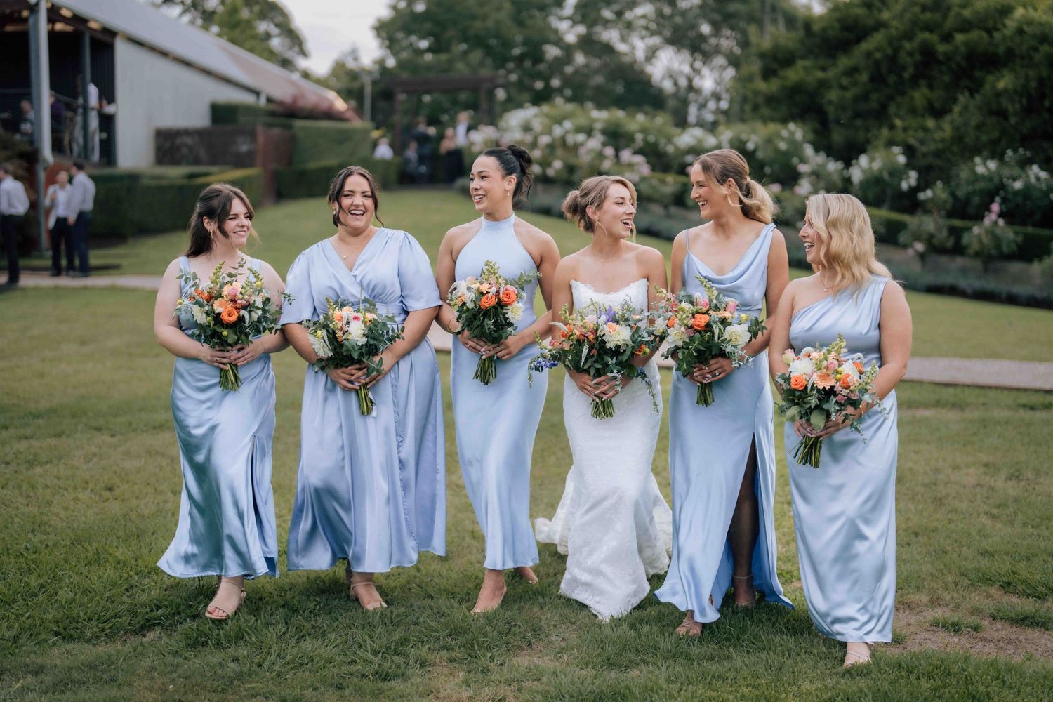 Five bridesmaids in light blue satin dresses walk together holding colorful bouquets at outdoor wedding.