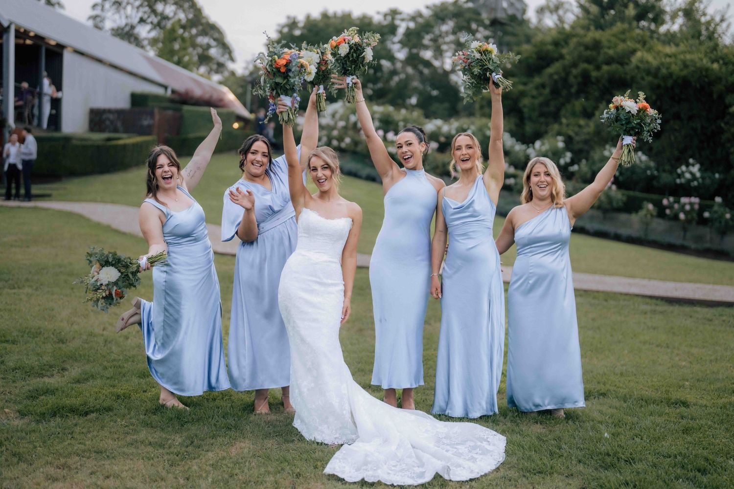 Bride and five bridesmaids in light blue dresses celebrate joyfully raising bouquets in lush garden wedding venue.