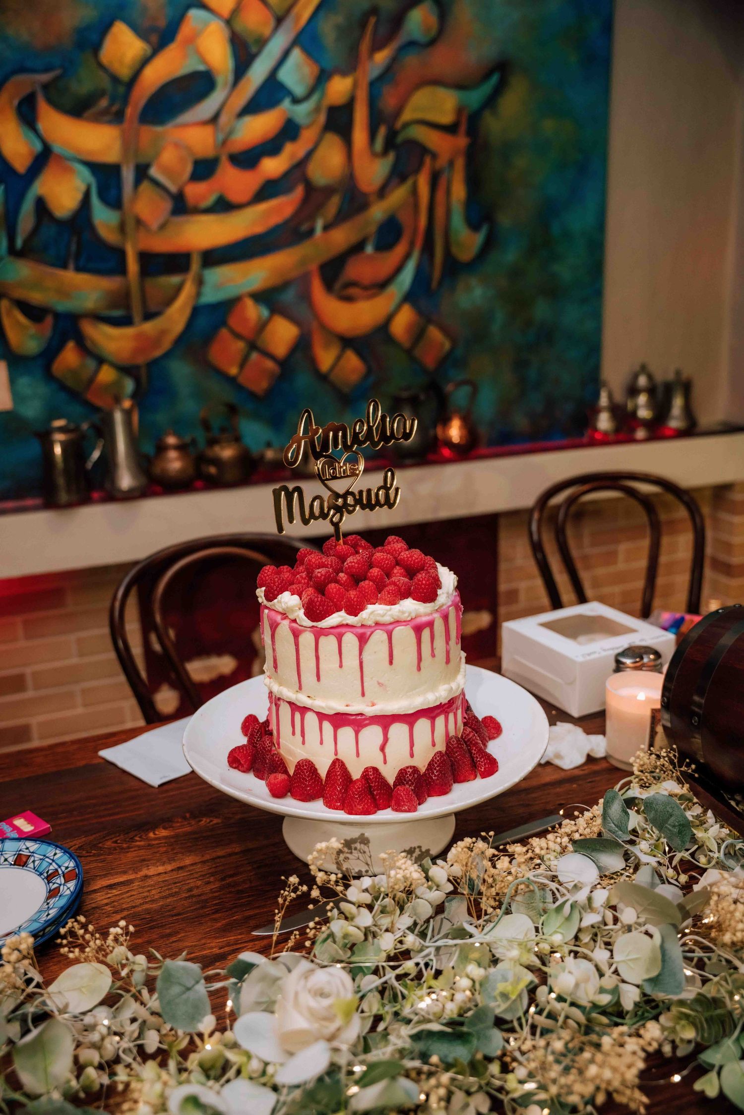 Two-tier white wedding cake with strawberries and gold topper reading Amelia and Masoud on decorated table.