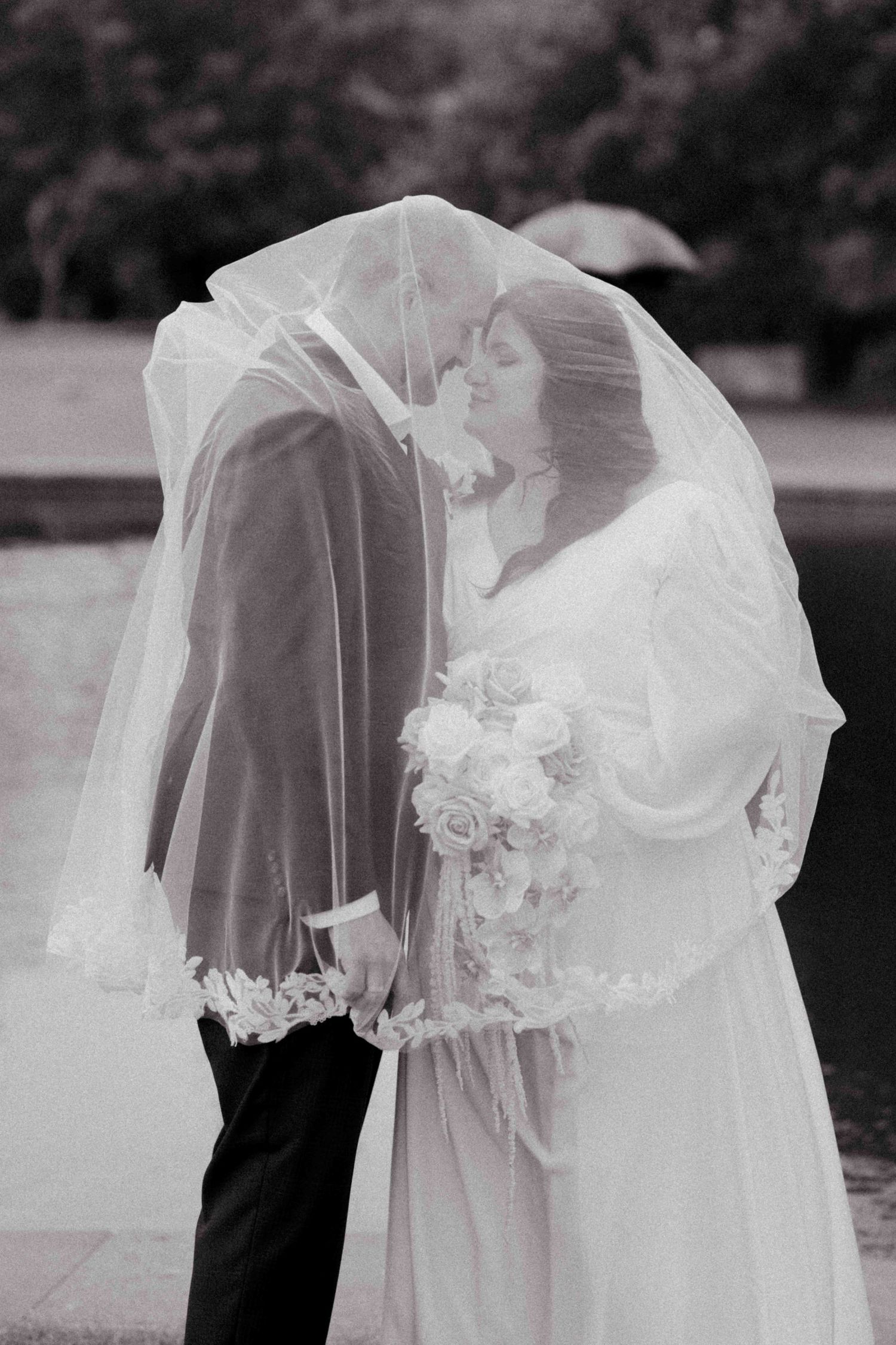 Black and white photo of bride and groom sharing a kiss under a flowing wedding veil, bride holding rose bouquet.