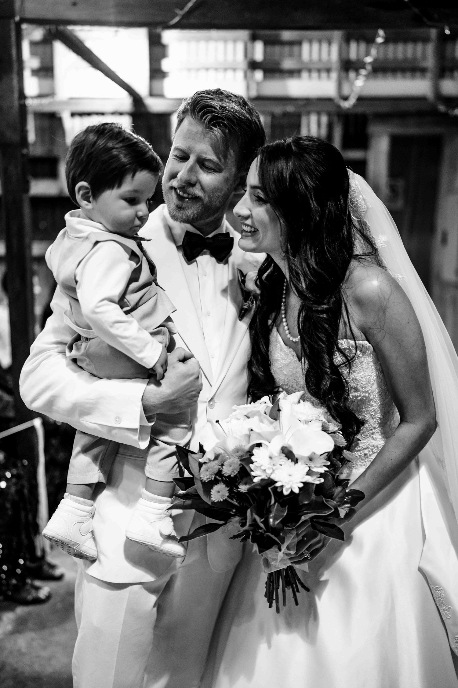Black and white wedding photo of bride and groom holding young boy in matching white suit, bride holds bouquet.
