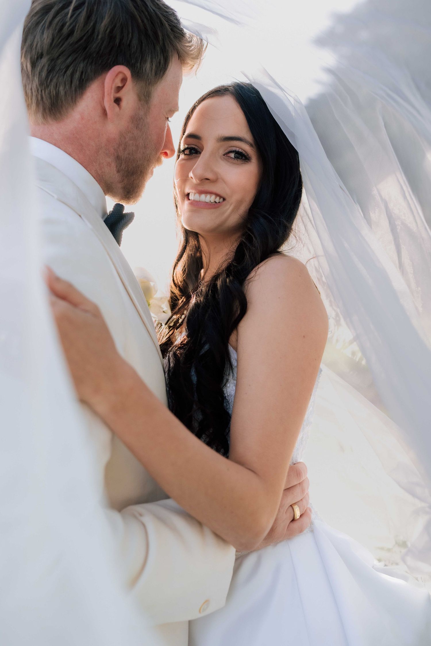 Smiling bride in white gown and veil embraces groom in white suit during outdoor wedding portrait.