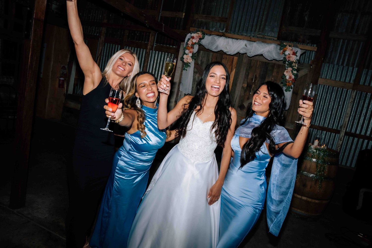 Bride posing with three bridesmaids in blue satin gowns, celebrating with drinks at wedding reception.