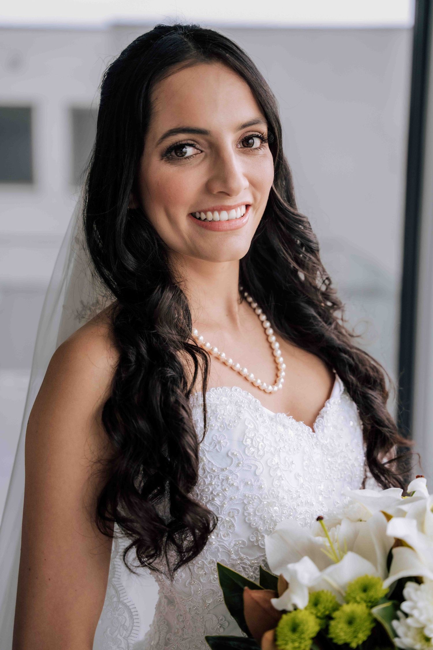 Smiling bride in white lace gown with pearl necklace holding white floral bouquet on wedding day.