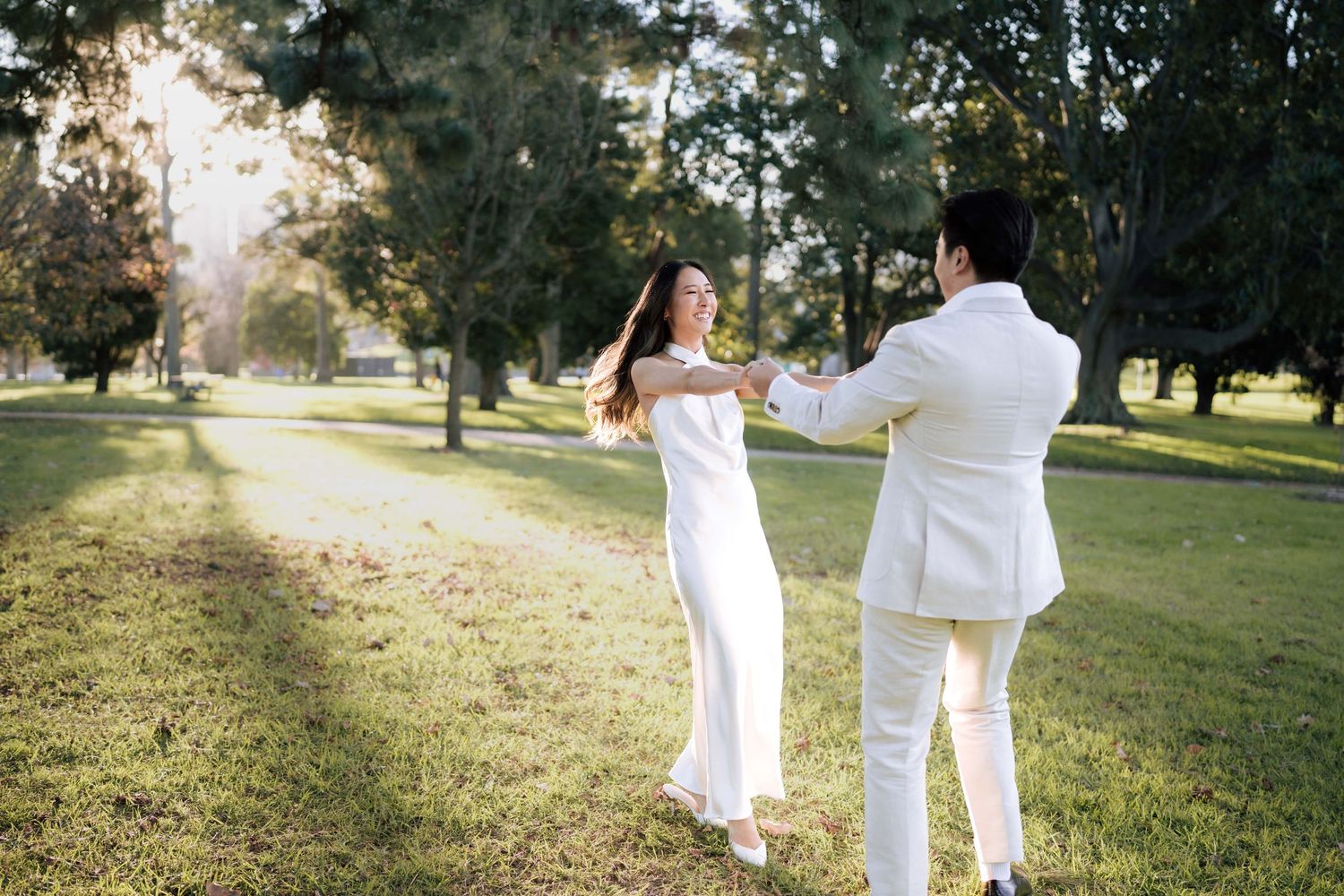 A couple in white outfits dancing joyfully in a sunlit park surrounded by lush green trees.