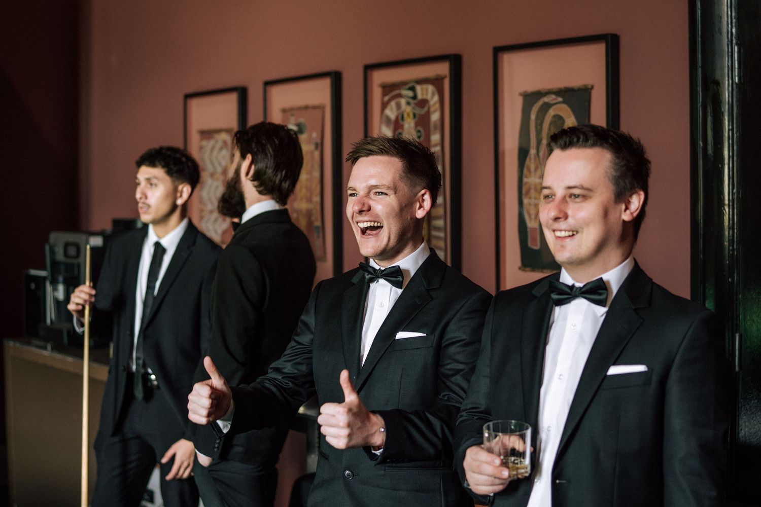 Men in black tuxedos smiling and giving thumbs up at a formal event, with framed artwork on the wall behind them.