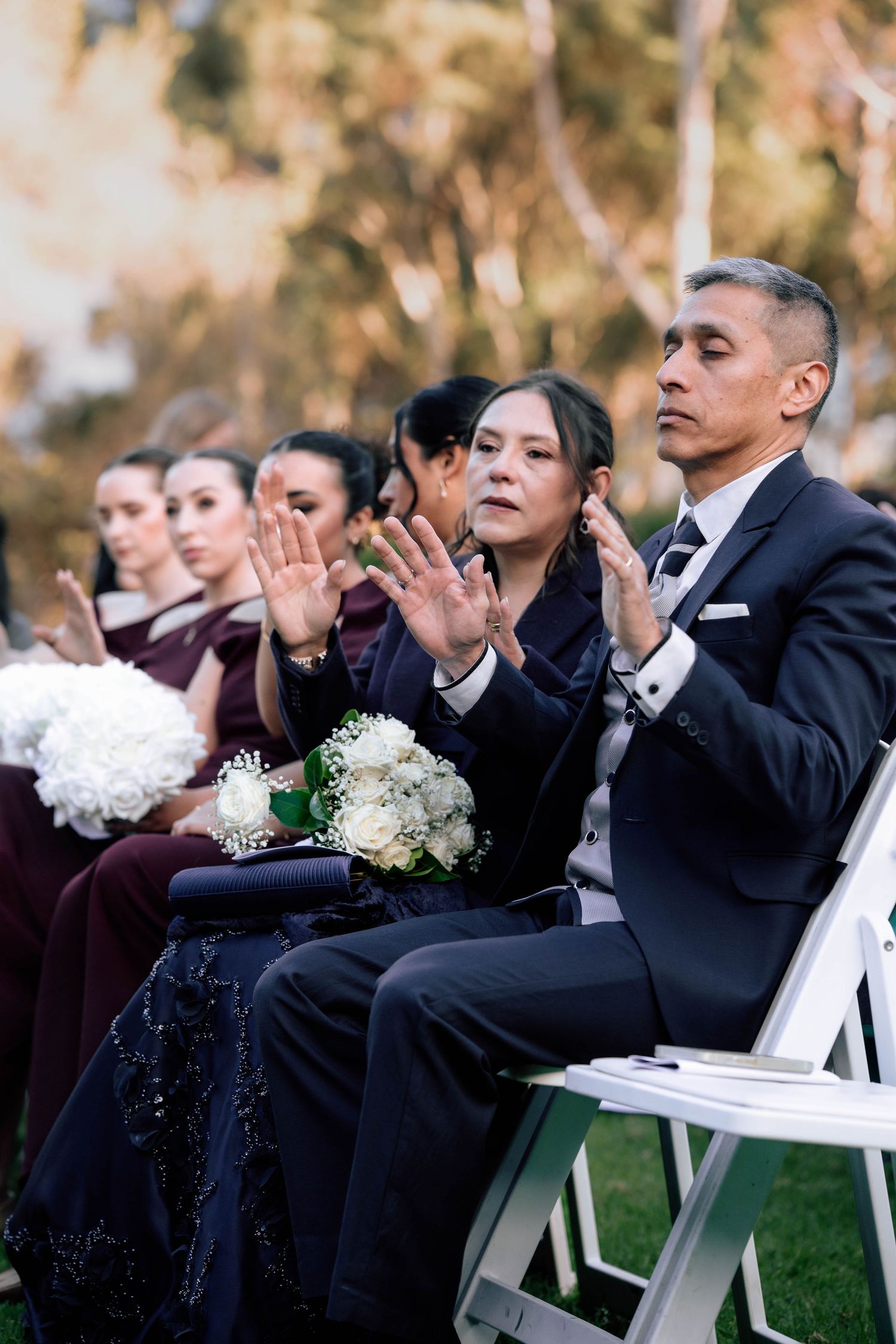 Wedding guests clap and celebrate during an outdoor ceremony with bridesmaids seated nearby.
