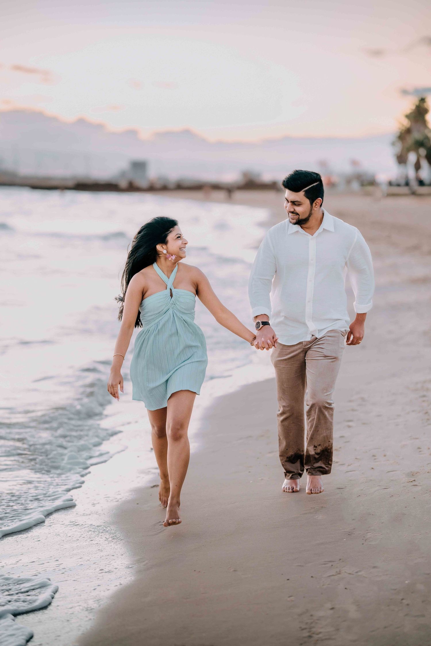 Happy Indian couple walking hand in hand along a beach at sunset smiling at each other.