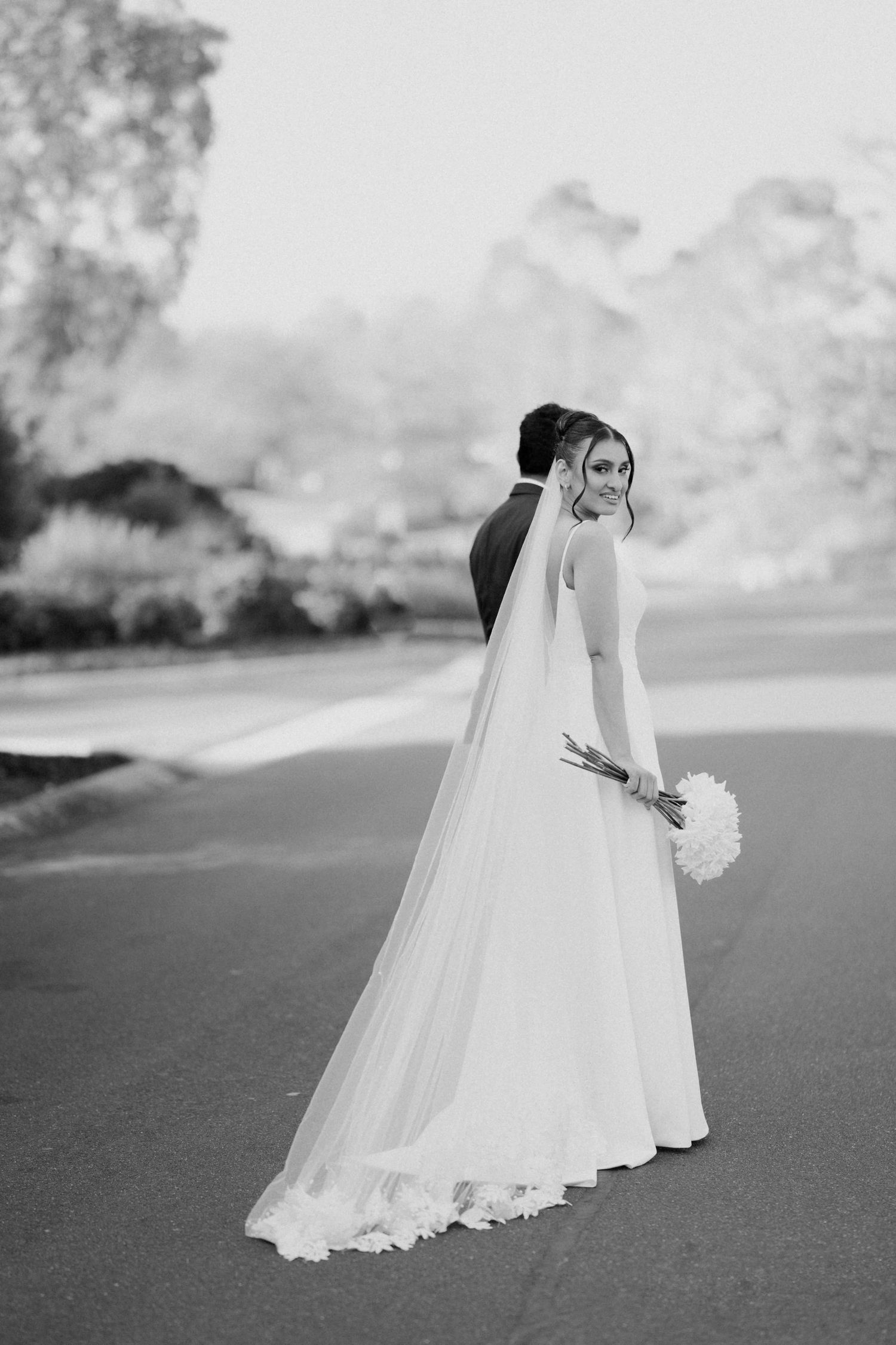 Black and white full-length portrait of bride in flowing gown with cathedral veil holding white bouquet.