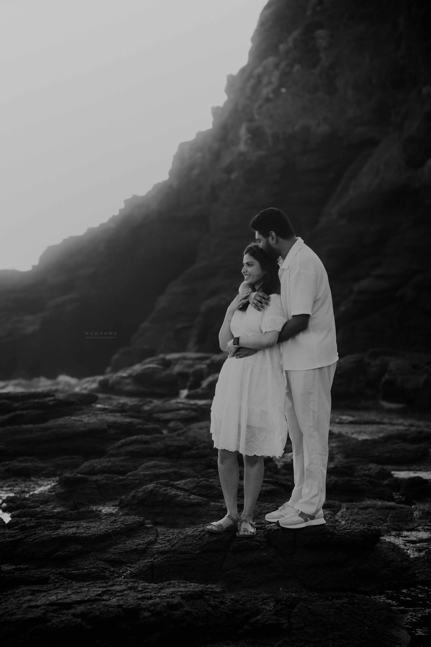 Black and white photo of couple in white standing together on rocky coastal cliffs.