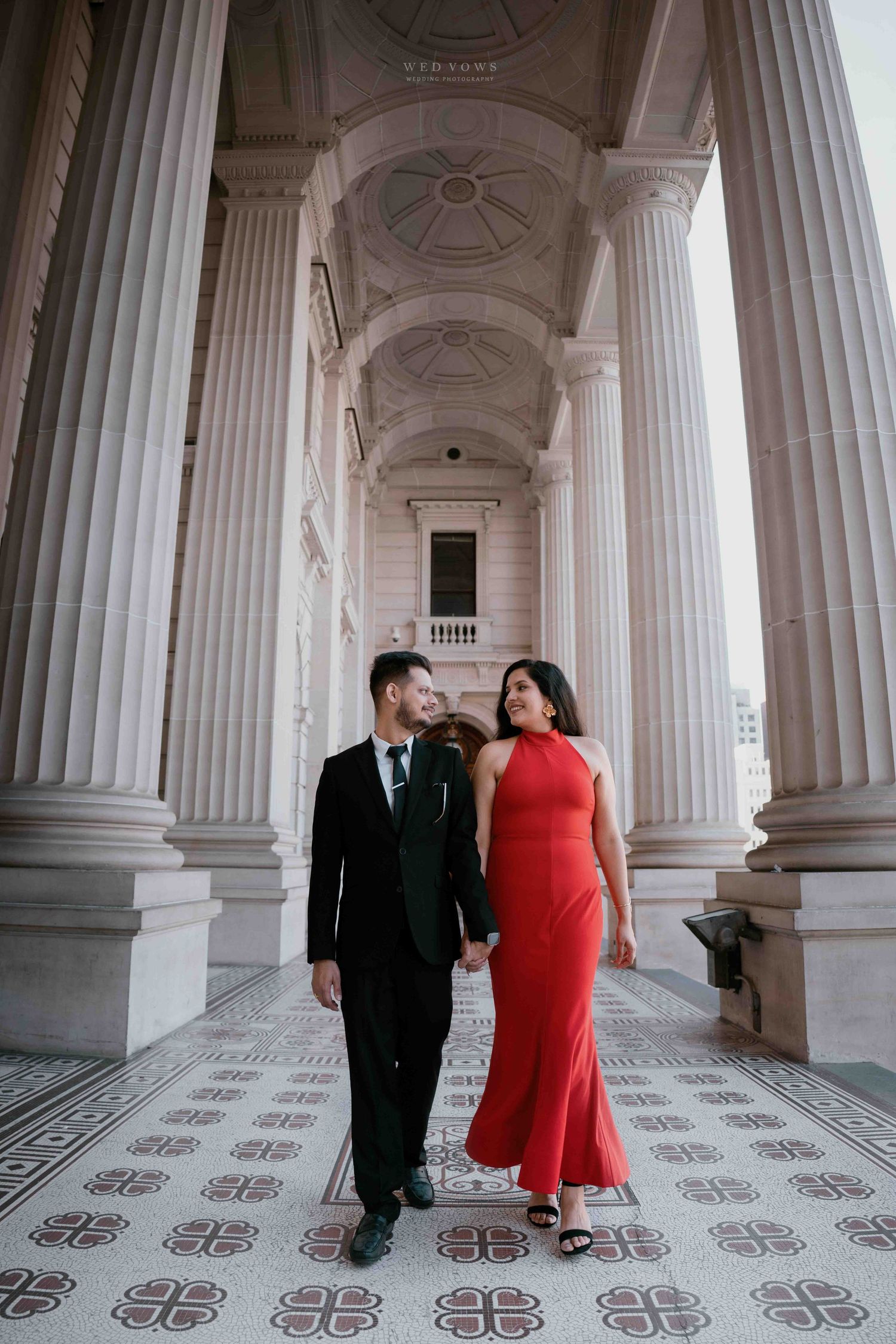 Couple walking hand in hand through grand white columned corridor, woman in red gown and man in black suit.