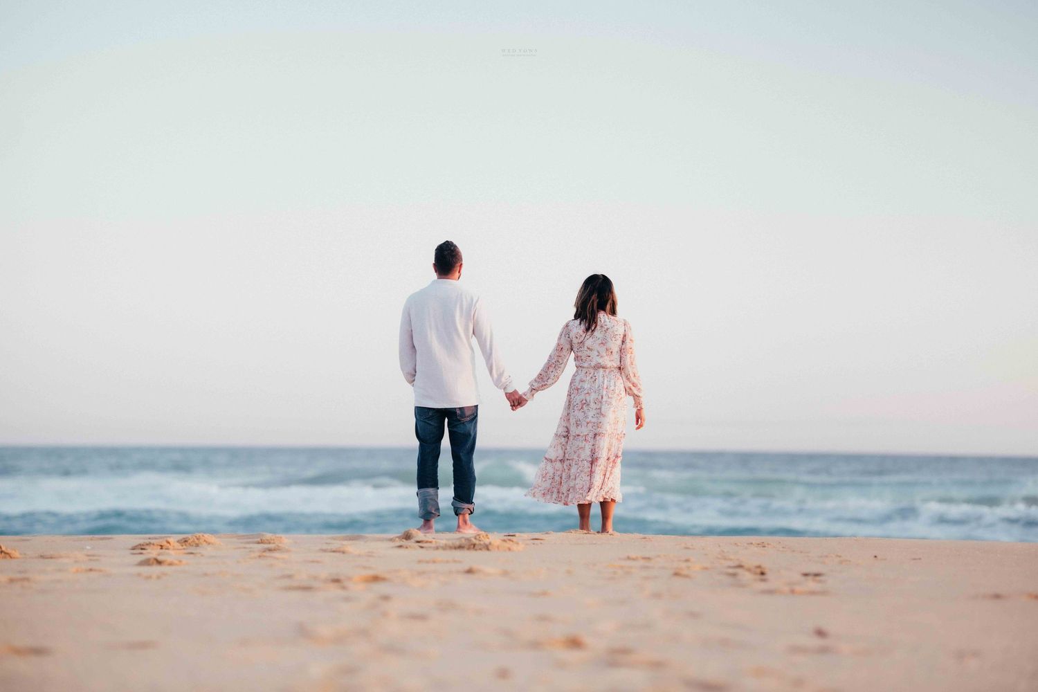 Romantic couple holding hands while standing barefoot on a sandy beach facing the ocean.