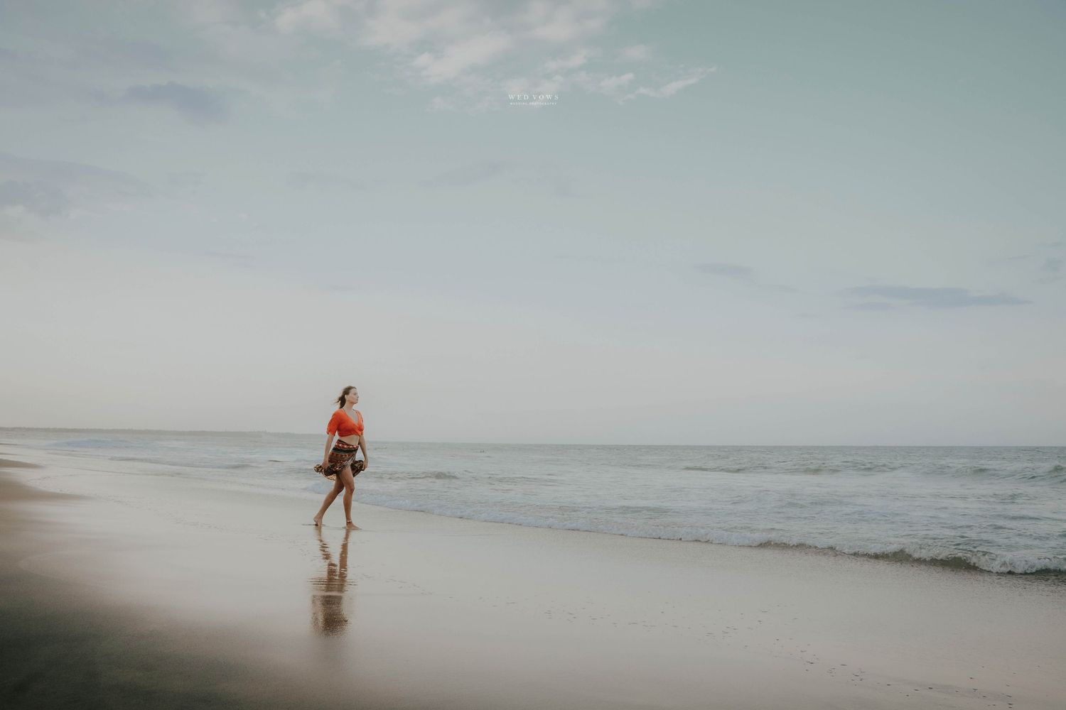 A woman in an orange top and patterned skirt walks alone along a serene beach with gentle ocean waves.