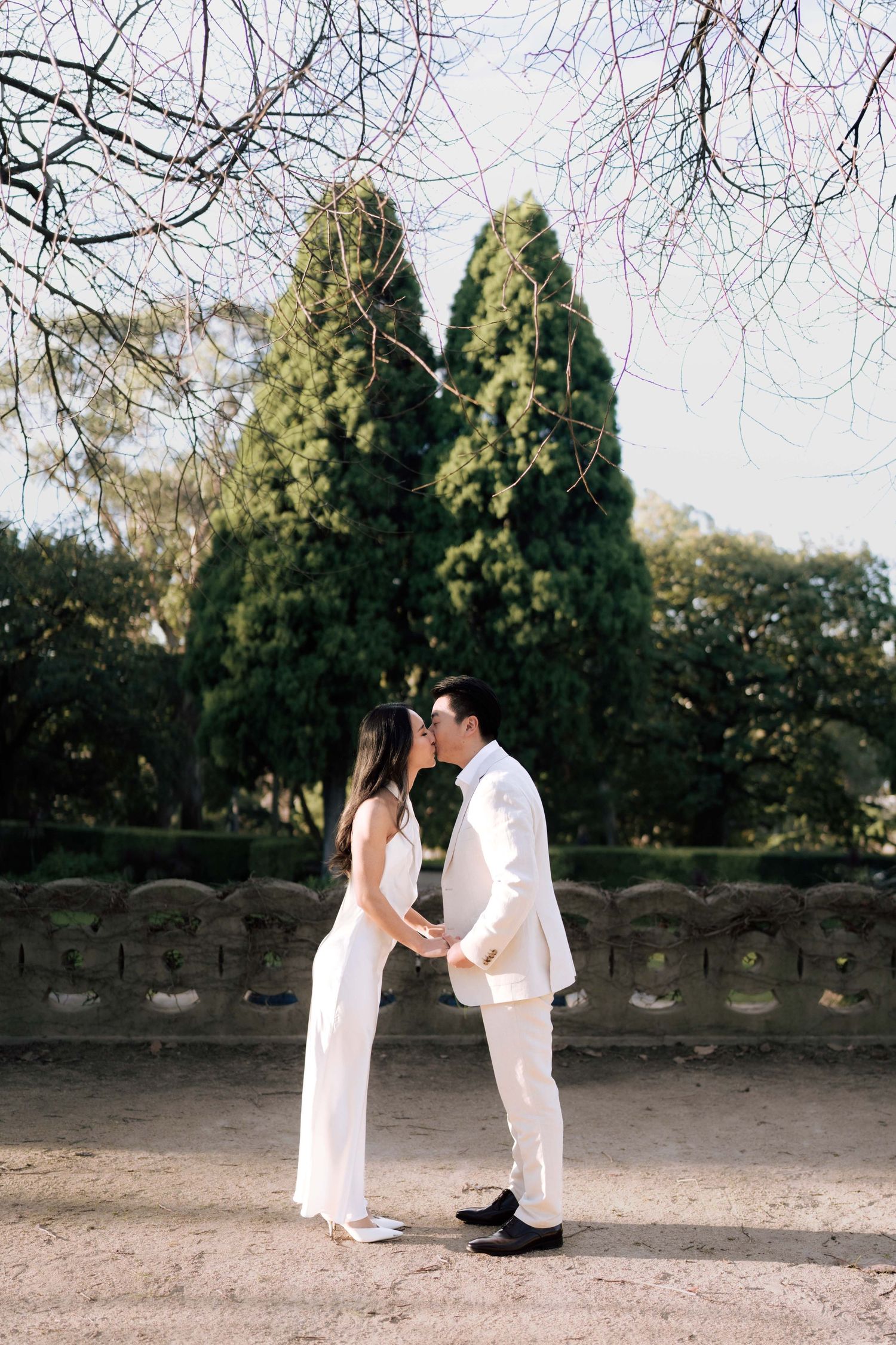 Couple in white attire sharing a romantic moment in a lush garden park with tall cypress trees.
