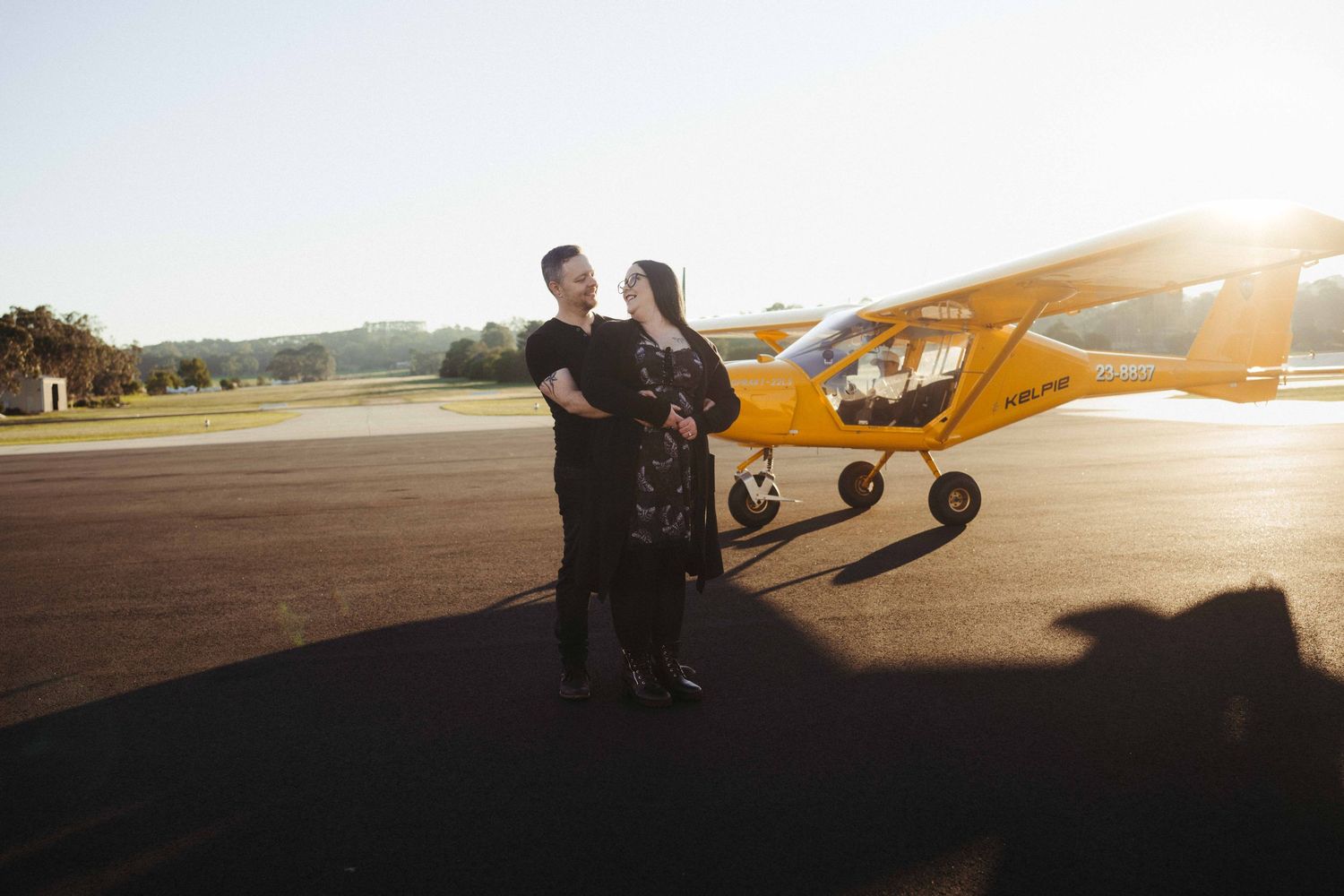 Couple embracing beside a yellow light aircraft on a sunny airstrip with golden backlight.