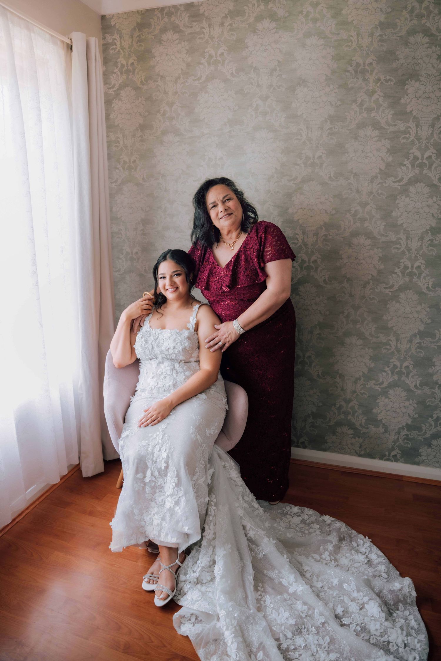 Bride in lace wedding gown poses with mother in burgundy dress against ornate wallpaper backdrop.