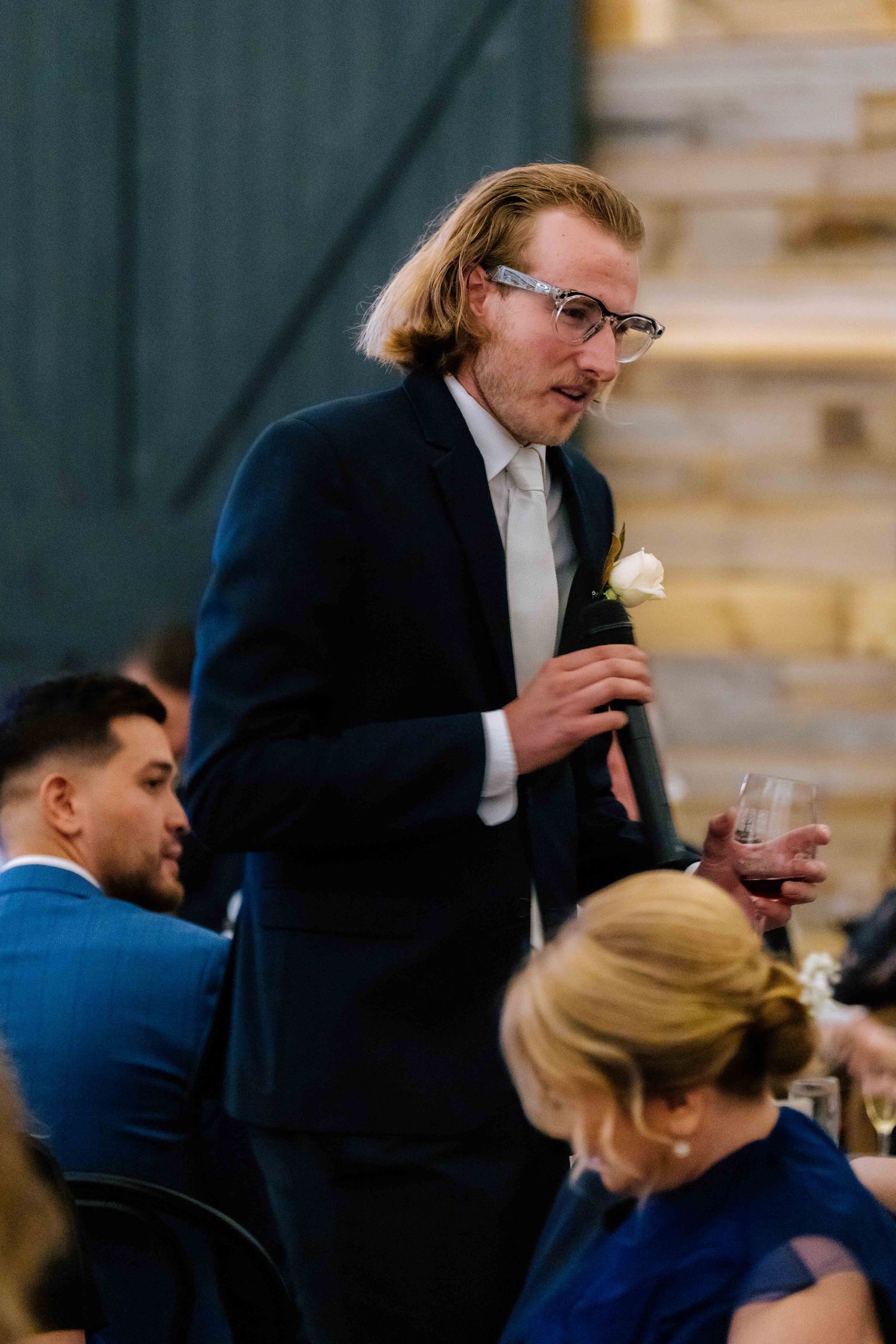 Young man in navy suit delivers wedding toast holding red wine glass at reception.