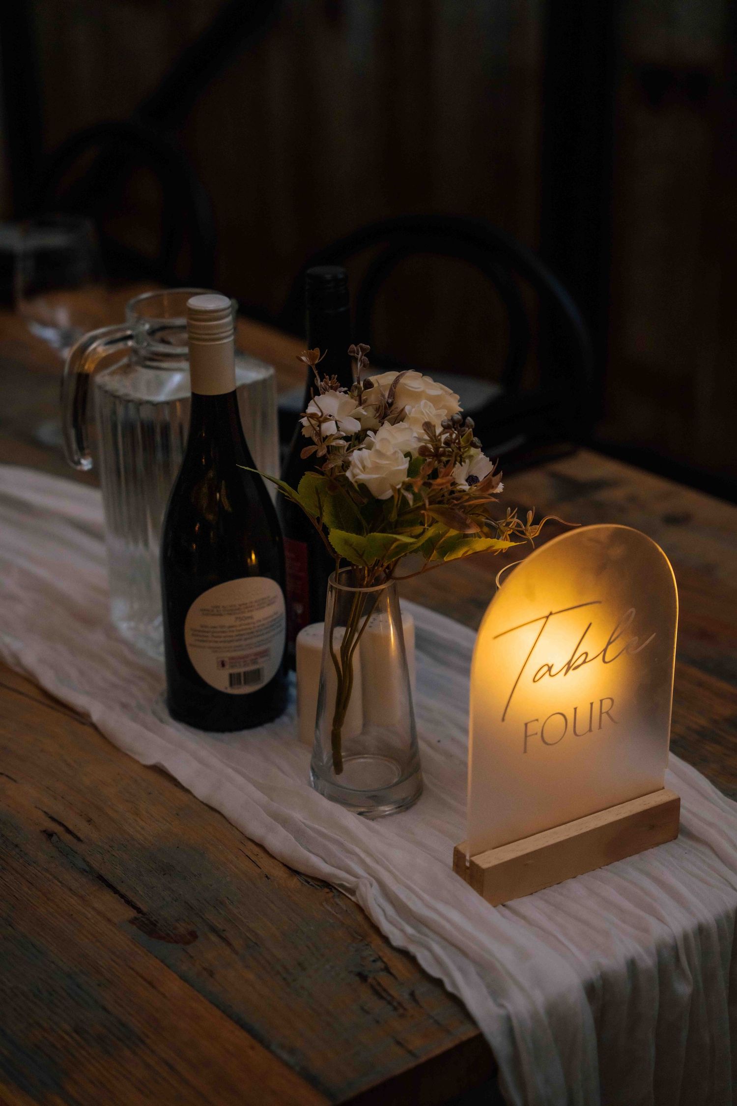 Glowing Table Four sign beside wine bottles and floral centerpiece on rustic wooden table.