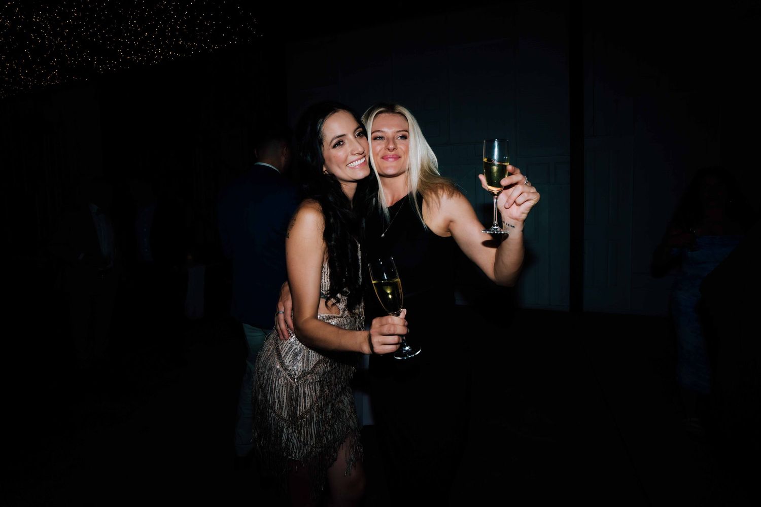 Two women in elegant dresses toasting with champagne glasses at a dark wedding reception.