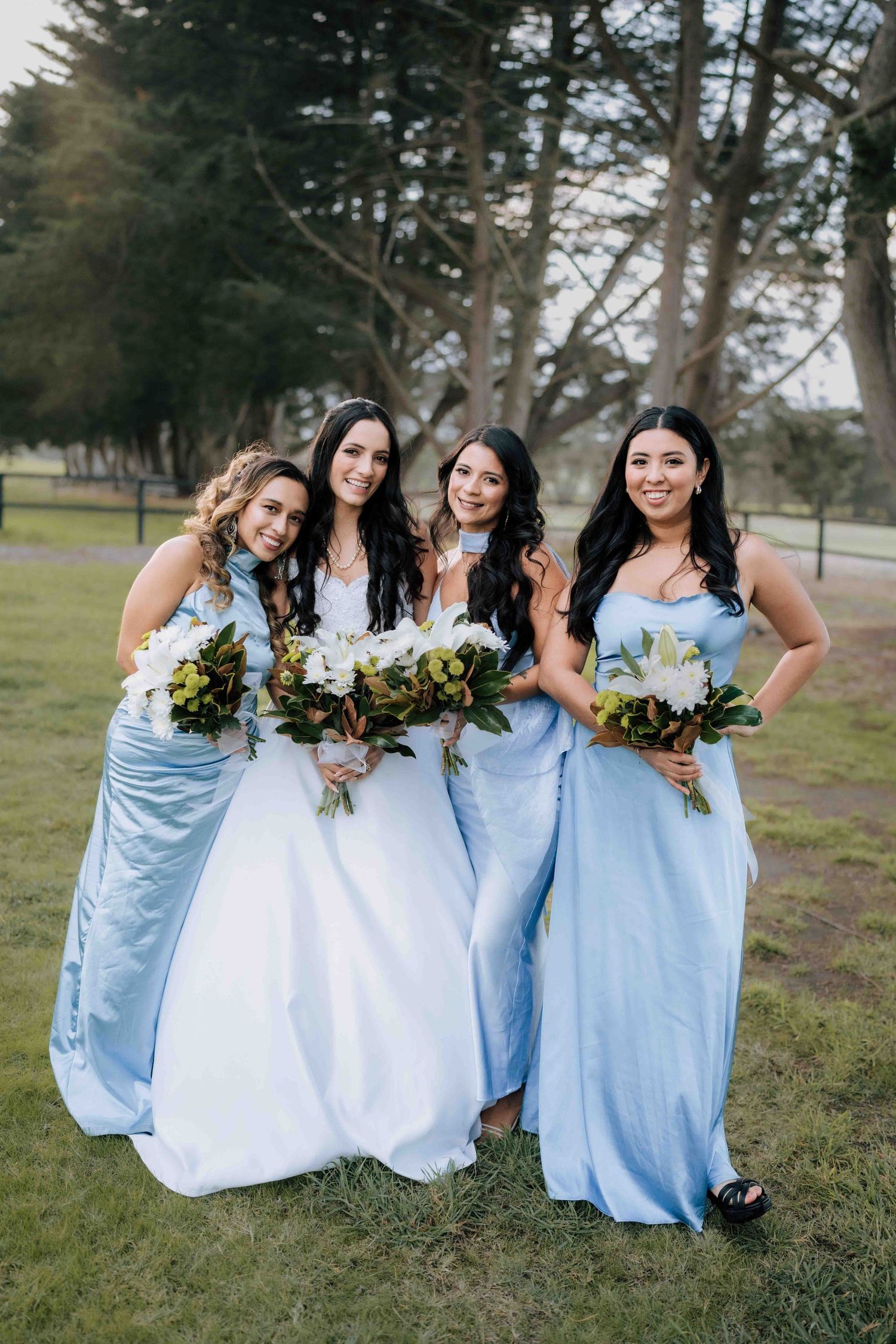 Four bridesmaids in light blue satin dresses holding white and green bouquets pose together outdoors at a wedding.