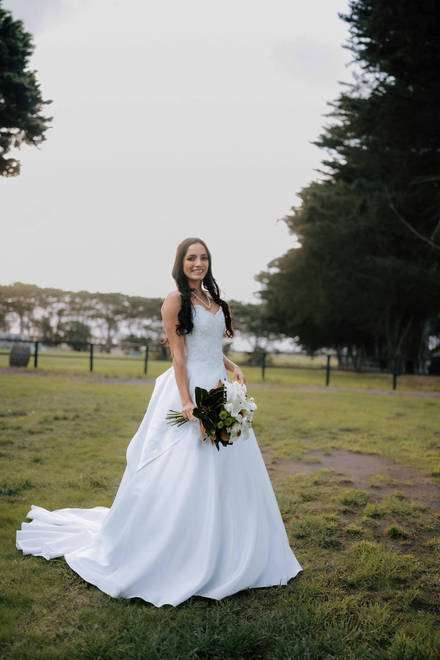 Bride in white ball gown holding bouquet stands on green grass with trees in background.