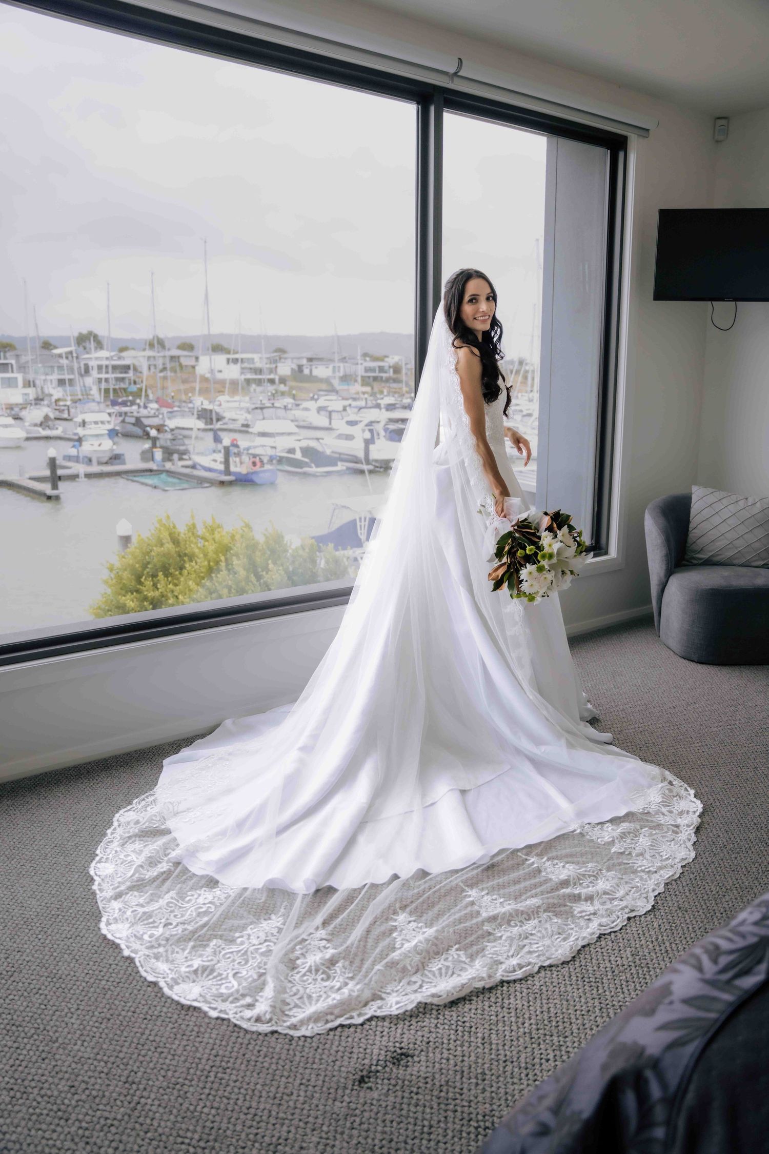 Bride in stunning white gown with lace train poses by large window overlooking a marina.